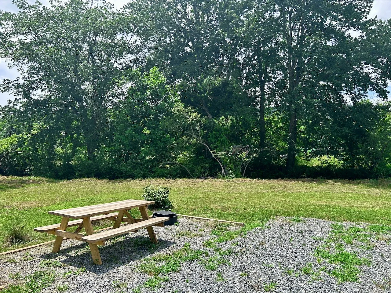 A wooden picnic table on a gravel surface, with a grassy lawn and large green trees in the background.