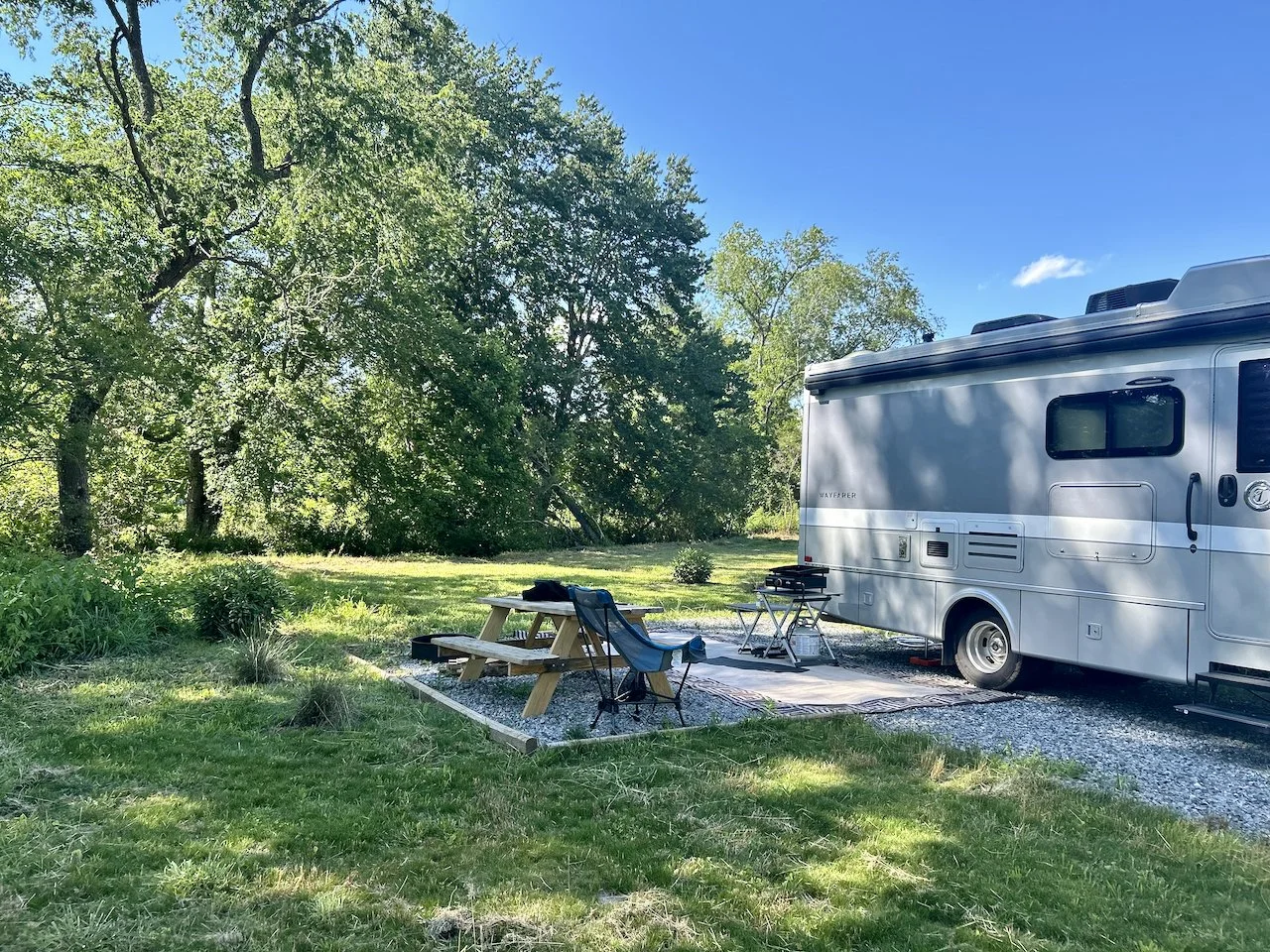 A camping site with a large silver RV, a picnic table, folding chairs, a small grill, and a mat on grass, surrounded by trees under a clear blue sky.