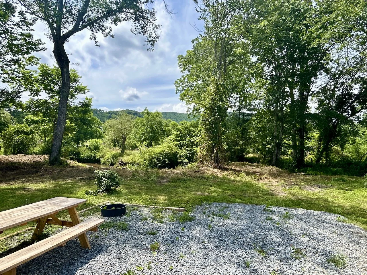 A scenic outdoor area with a gravel patio, a wooden picnic table, a black round planter, lush green trees, and a hill in the background under a partly cloudy sky.