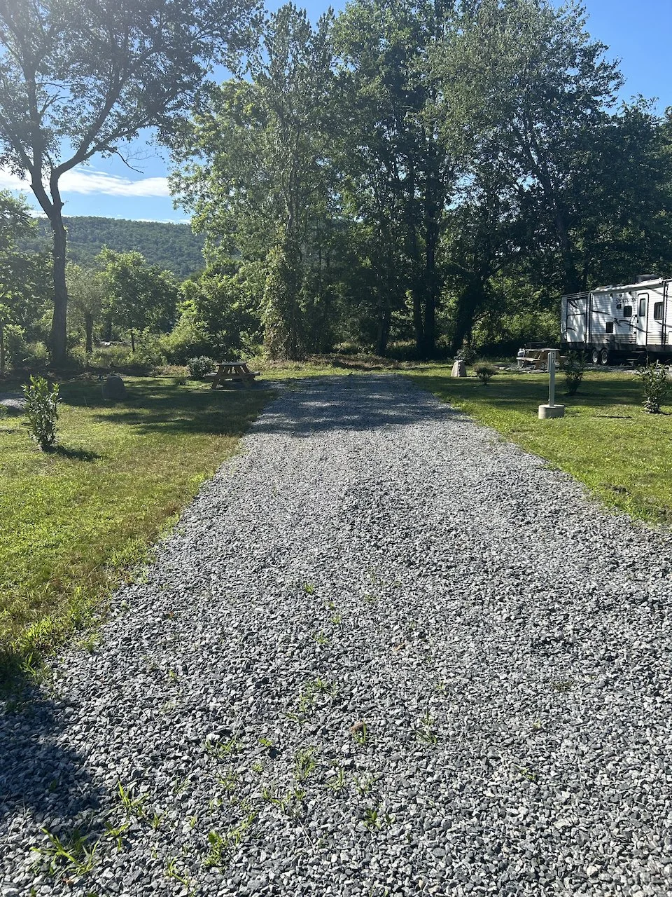 Gravel pathway leading through a grassy area with trees, a picnic table, a white RV, and camping equipment under a clear blue sky.