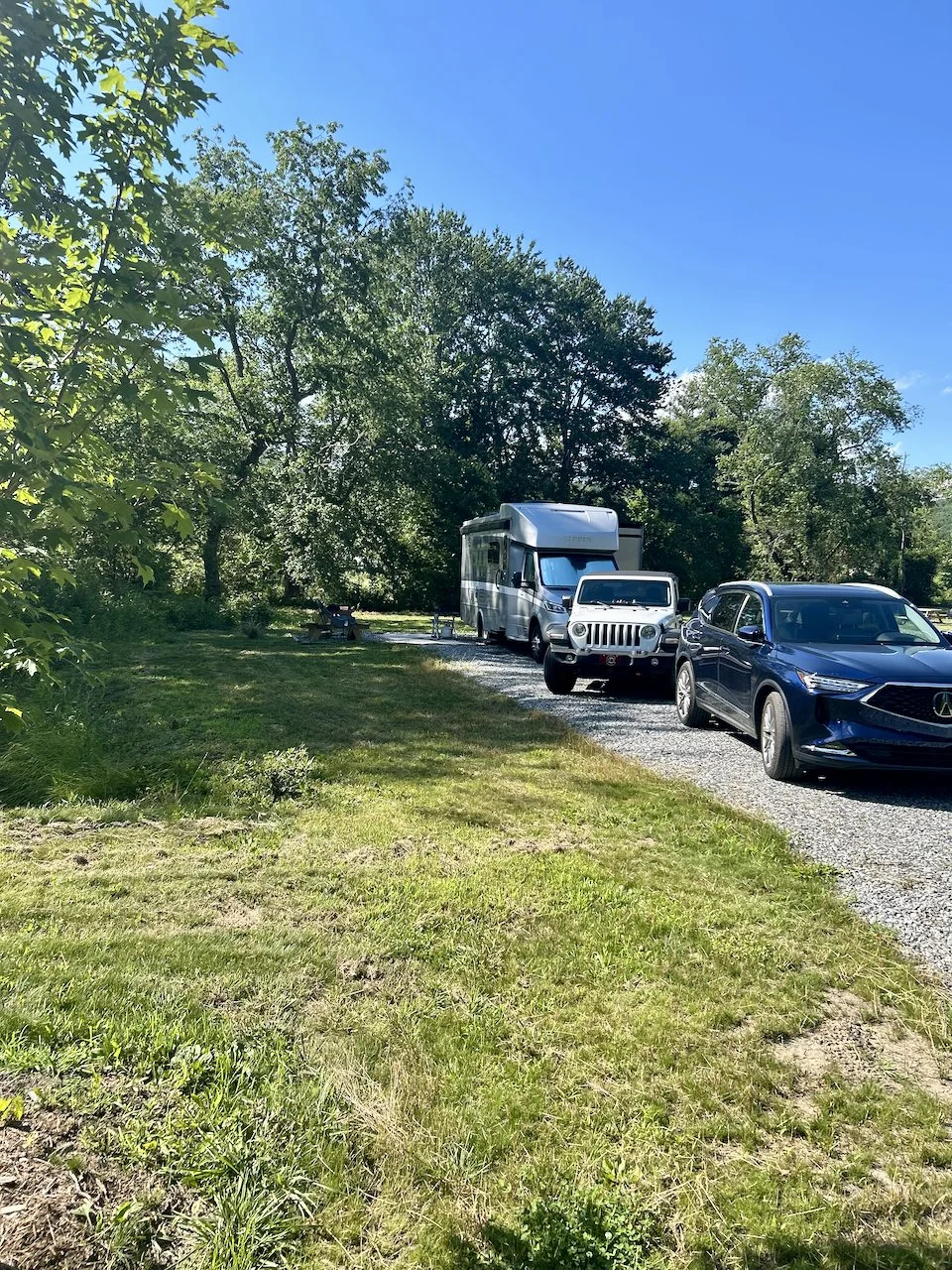 A gravel road with three parked vehicles: a recreational vehicle, a white Jeep, and a black SUV, surrounded by green trees and grass under a clear blue sky.