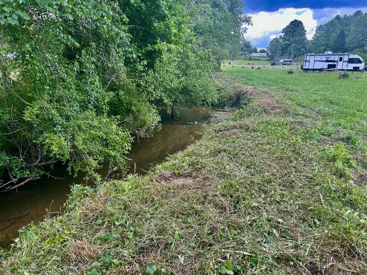 A small creek runs through a grassy area with dense green bushes on the left. In the background, there is an open field with an RV and trees, under a partly cloudy sky.
