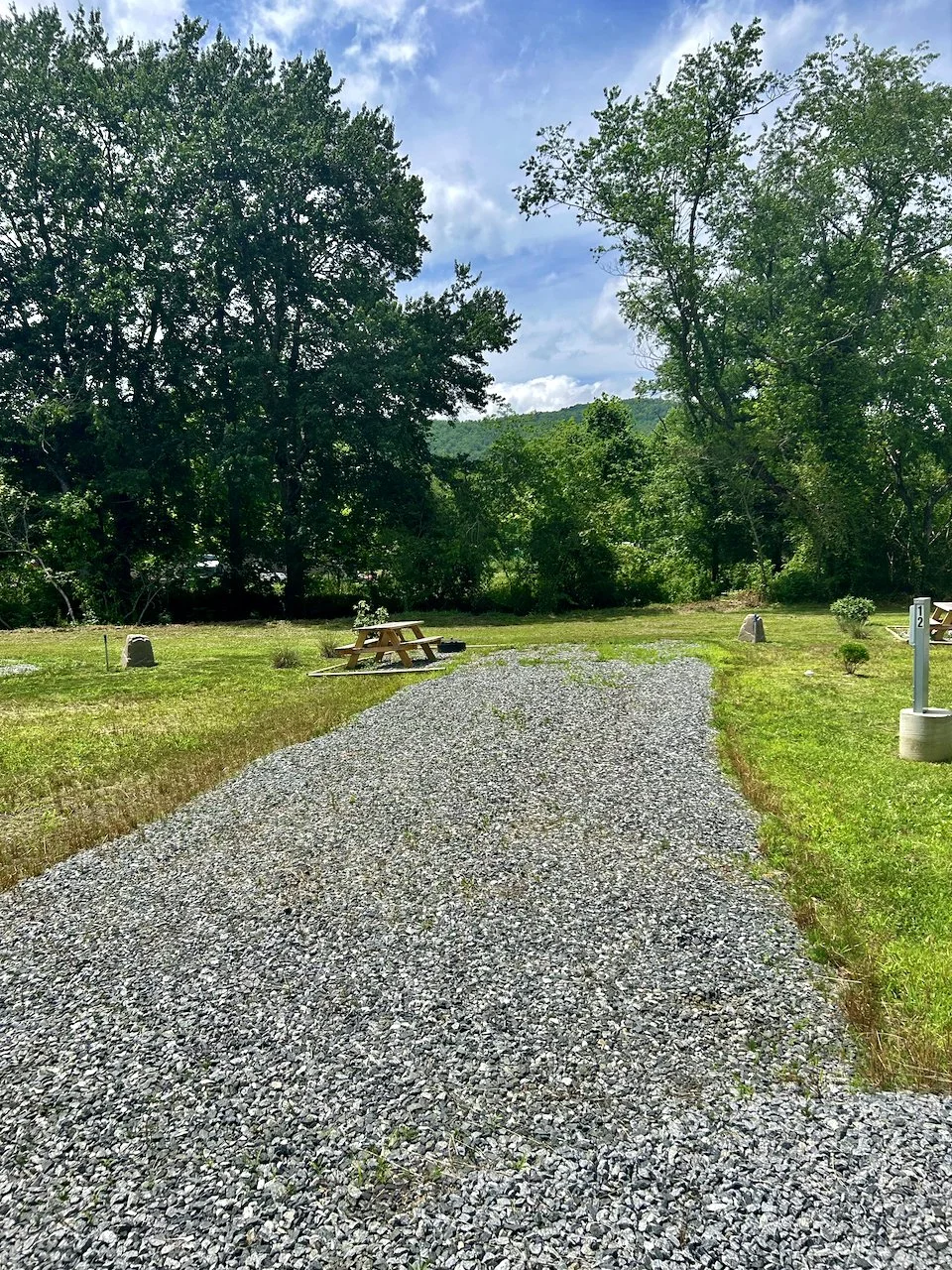 Gravel pathway leading into a grassy clearing with trees in the background, a picnic table on the left, a small shrub on the right, and a blue sky with some clouds.