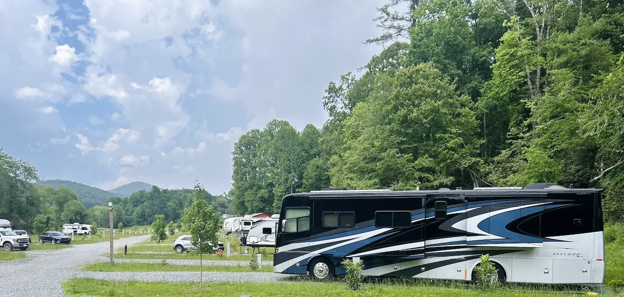 A large black, white, and blue RV parked in a campsite with other RVs and vehicles around. The campsite is surrounded by green trees and hills under a partly cloudy sky.