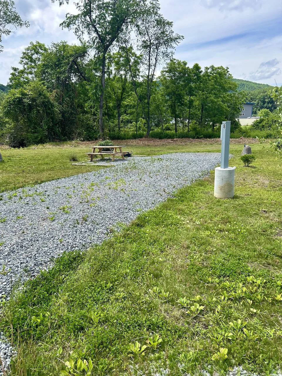 A gravel pathway in a grassy outdoor area with a picnic table, a signpost labeled '8', and trees in the background.