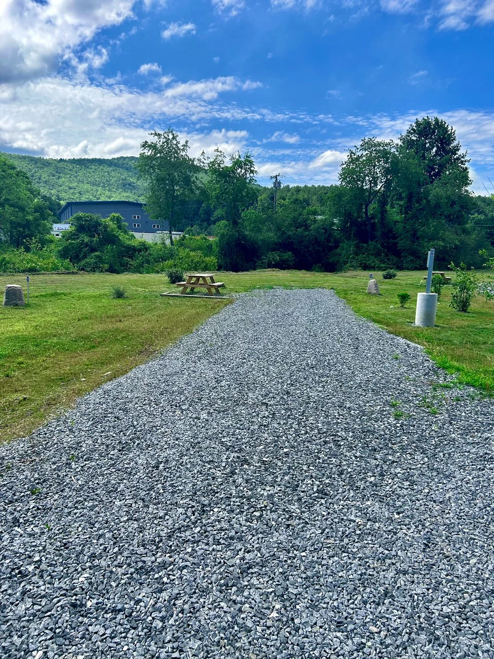 Gravel pathway leading to a grassy area with trees and mountains in the background, under a partly cloudy sky.