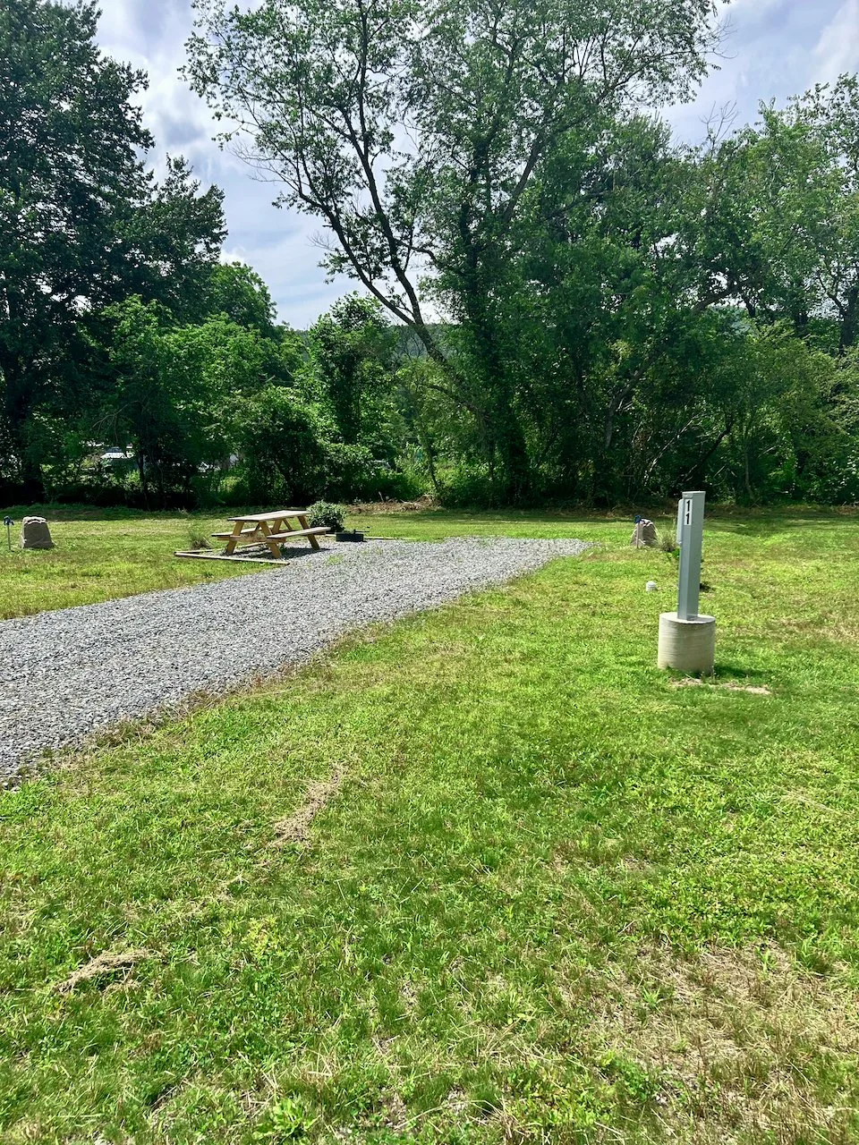 A grassy park area with a gravel pathway, a wooden picnic table, a small black object near the table, a metal utility pole on a concrete base, and surrounded by green trees under a partly cloudy sky.