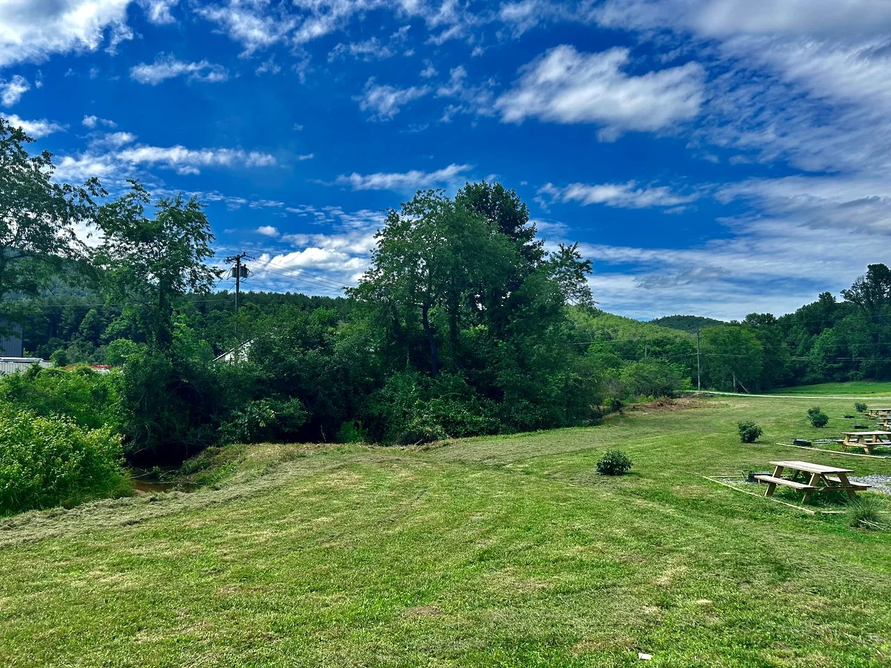 Picnic tables on a grassy hillside with trees, hills in the distance, and a partly cloudy sky.