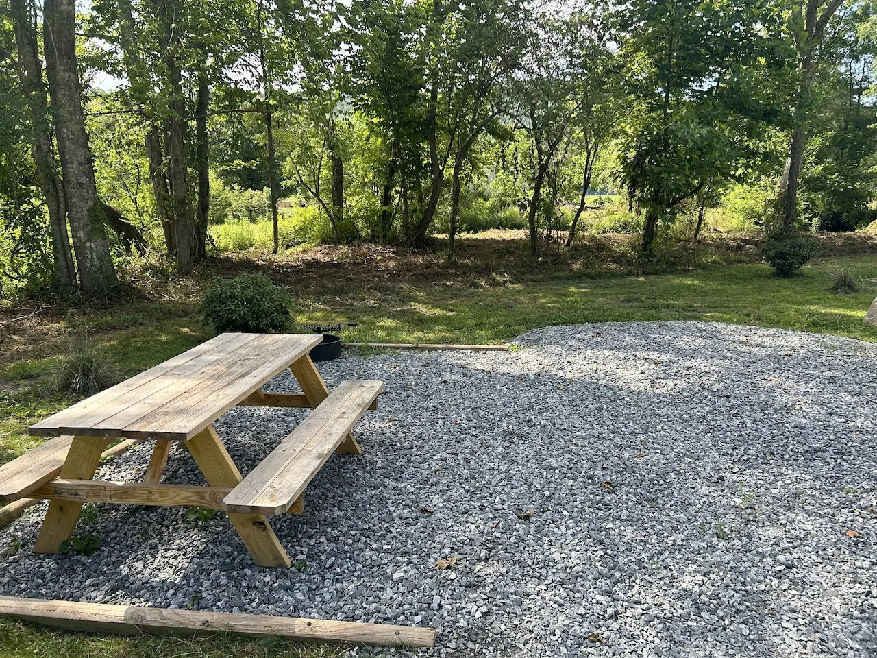 Wooden picnic table on a gravel surface in a grassy, wooded outdoor area with trees and sunlight filtering through the leaves.