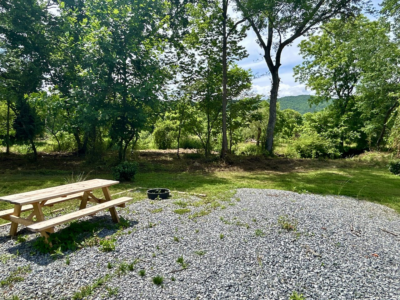 A backyard with a gravel area, wooden picnic table, black bucket, grassy patch, and trees with a mountain and cloudy sky in the background.