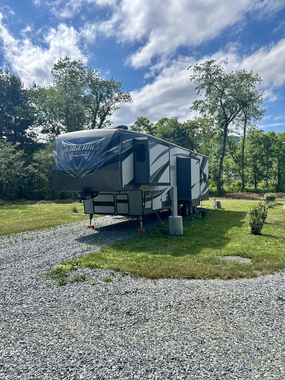 A camper trailer parked on a gravel pad with grassy surroundings and trees under a partly cloudy sky.