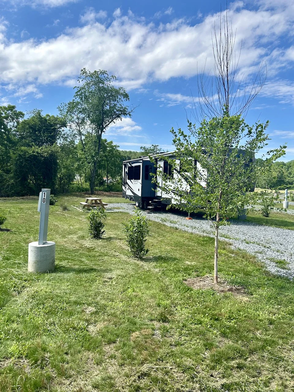 A white travel trailer parked on a gravel pad in a grassy area with trees and a picnic table, under a partly cloudy blue sky.