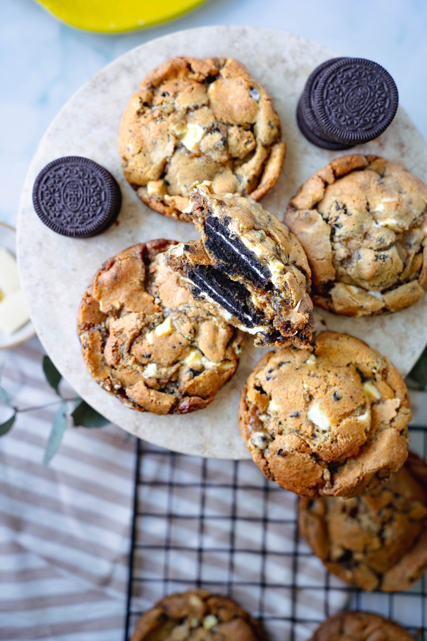 Bildet viser en rund marmorplate med fire store cookies. En av cookiene er delt i to og viser fyll av Oreo-kjeks og hvit sjokolade. Rundt cookiesene ligger to hele Oreo-kjeks. I bakgrunnen ser man en skål med hvit sjokolade, en sitron på et gult fat