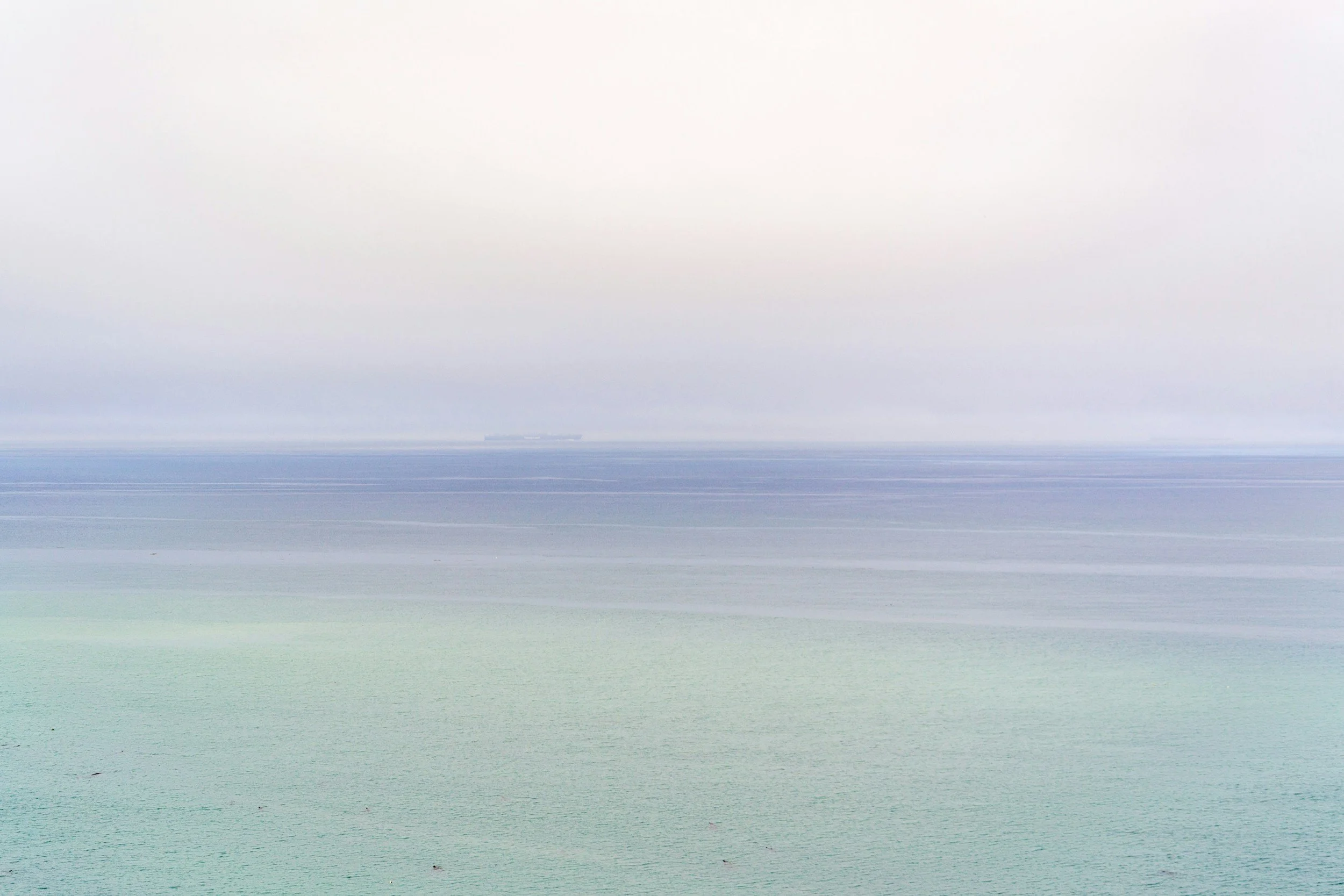 A misty seascape with calm, muted-colored water and a distant, barely visible ship on the horizon.