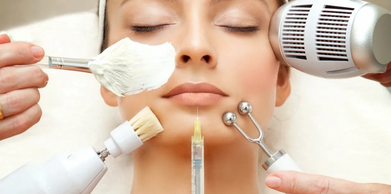 A woman receiving a facial treatment with various skincare tools, including a brush, a syringe, a roller, a fan, and a spatula, near her face with closed eyes.