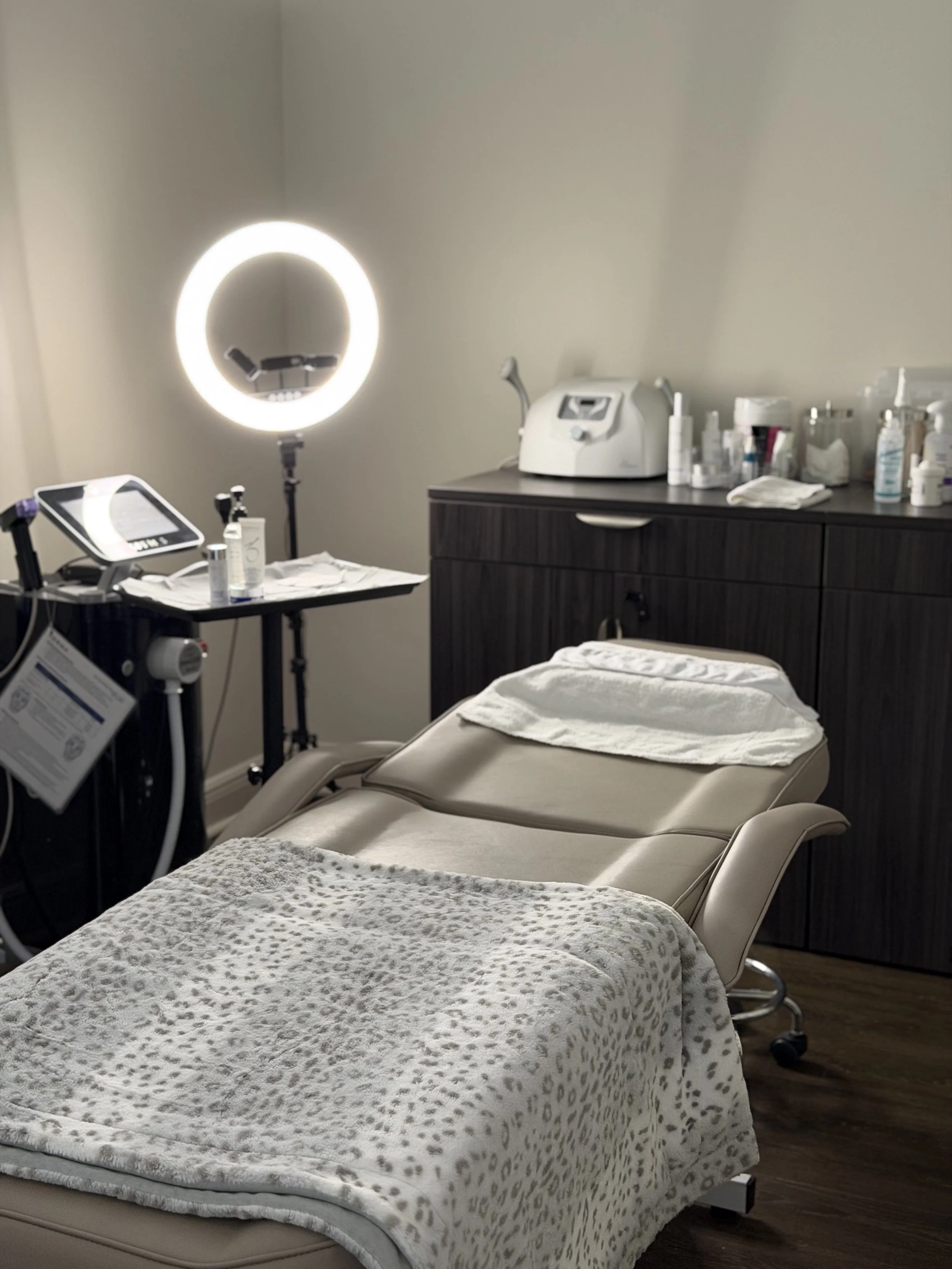 Medical treatment room with a treatment bed covered with a leopard print blanket and a towel, medical equipment including a ring light, and a cabinet with various supplies.