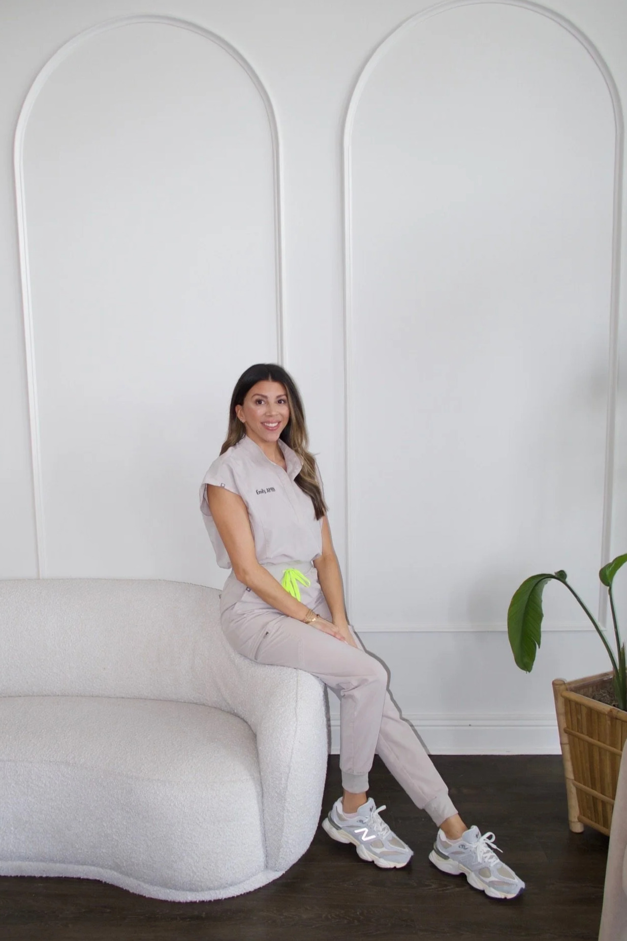 A woman with long wavy hair sitting on a light-colored armchair against a white wall, smiling at the camera.