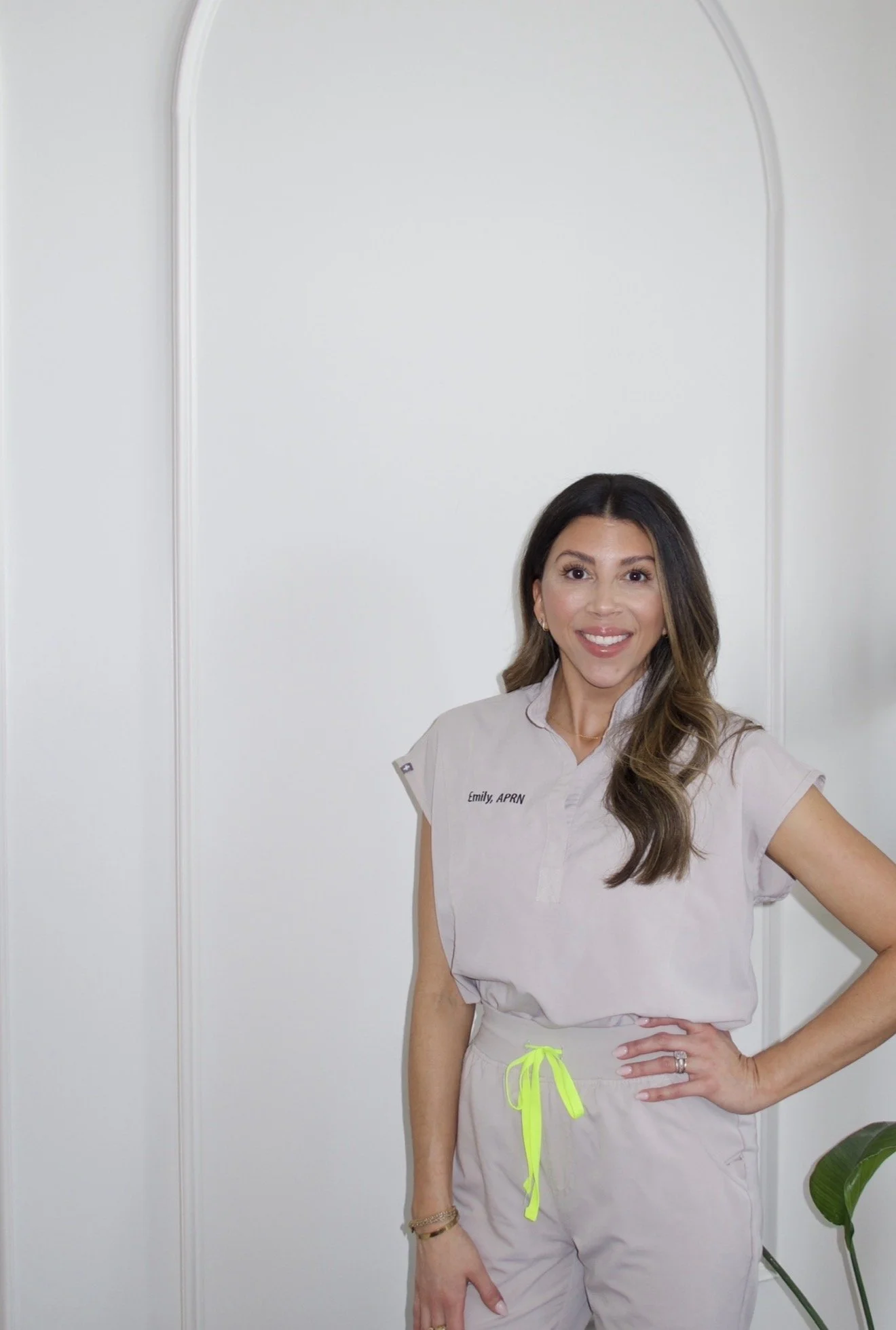 A woman with long wavy hair sitting on a light-colored armchair against a white wall, smiling at the camera.