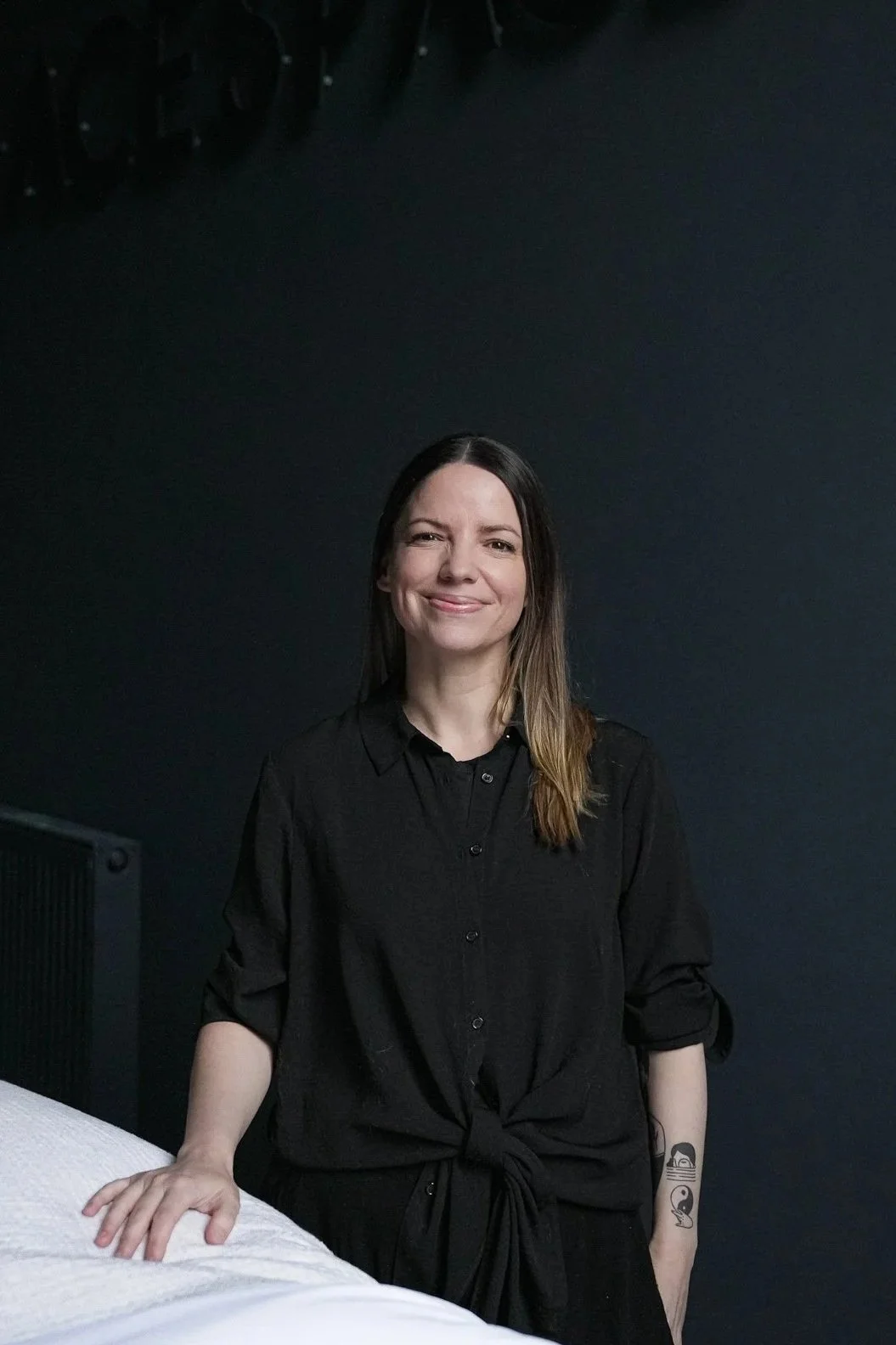 A woman with long hair, wearing a black shirt, smiling and standing in front of a dark background.
