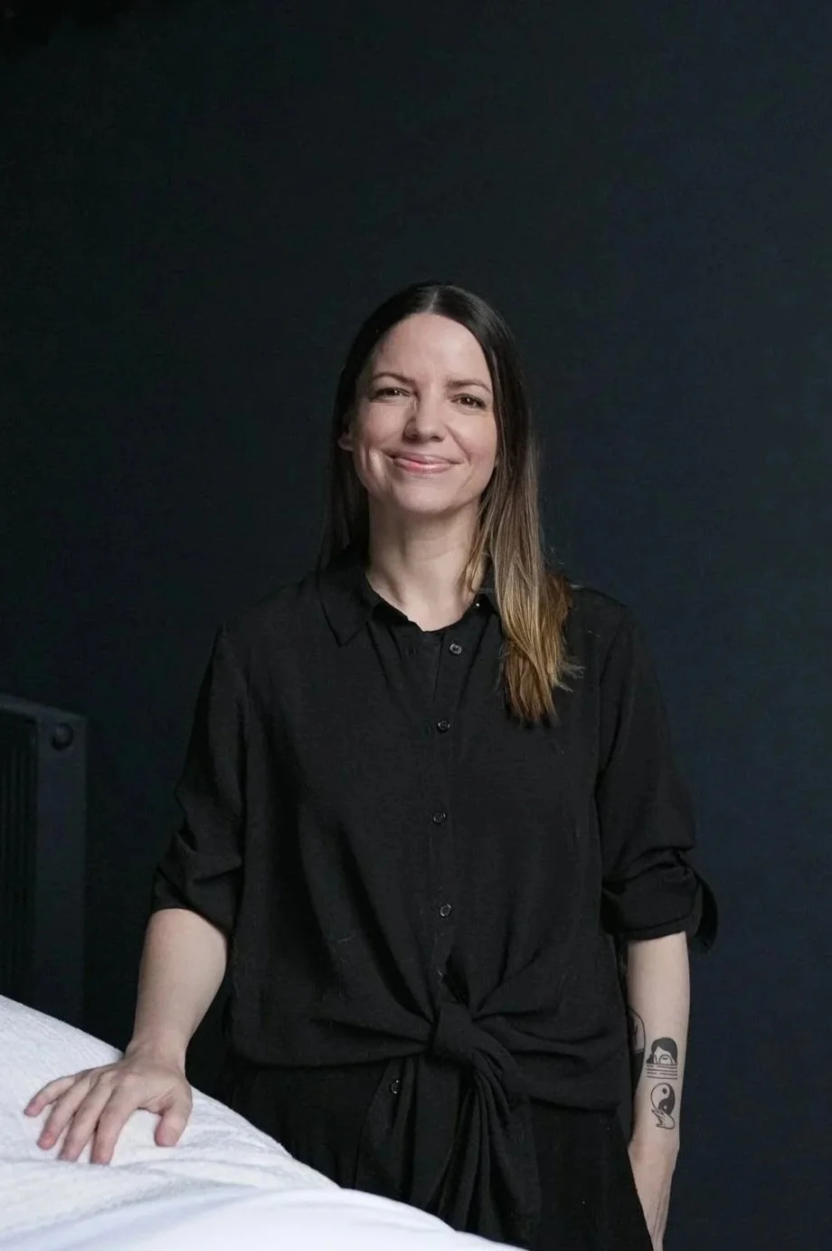 A woman in a black shirt with rolled-up sleeves, standing against a dark background, smiling with her left hand resting on a white bed or surface, and showing two tattoos on her right arm: a girl with goggles and a small whale.