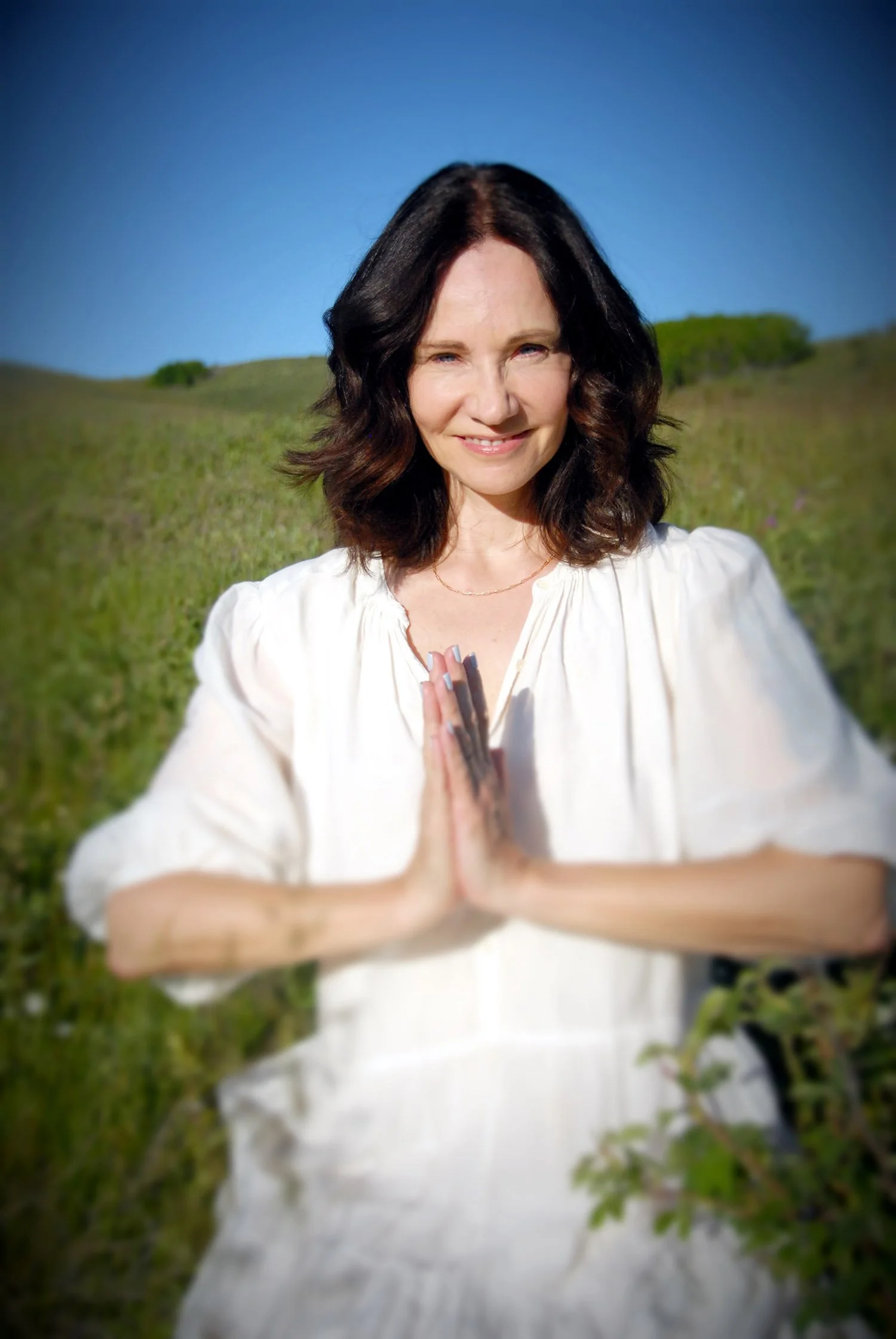 Victoria Brady sitting in a field against a bright blue sky, greenery, and hills in the background. She is smiling, wearing a white dress, and has her hands in a prayer position, anjali mudra.