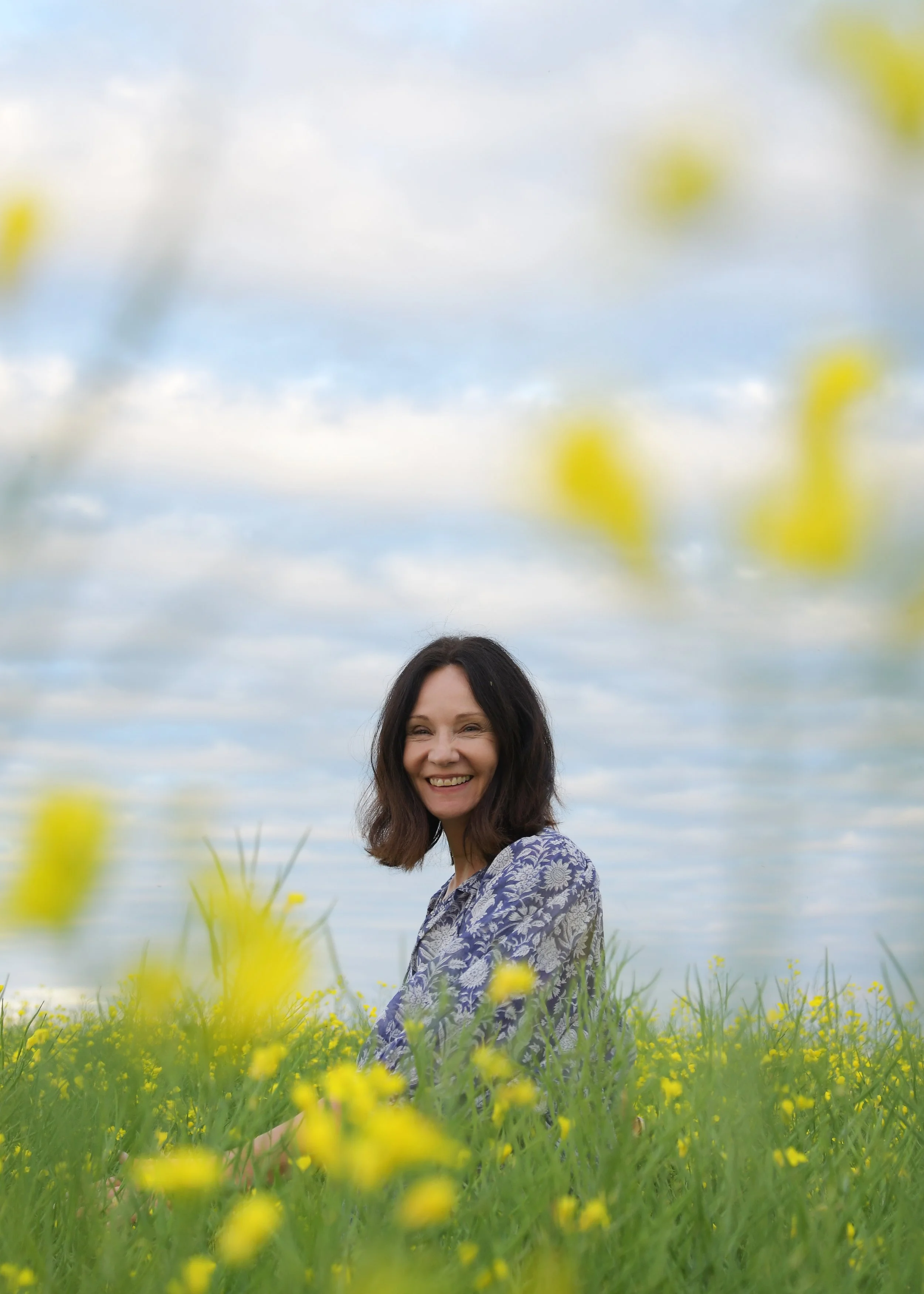 A woman (Victoria Brady) with dark, shoulder-length hair and a floral shirt smiling in a field of yellow flowers with a blue sky and clouds in the background.