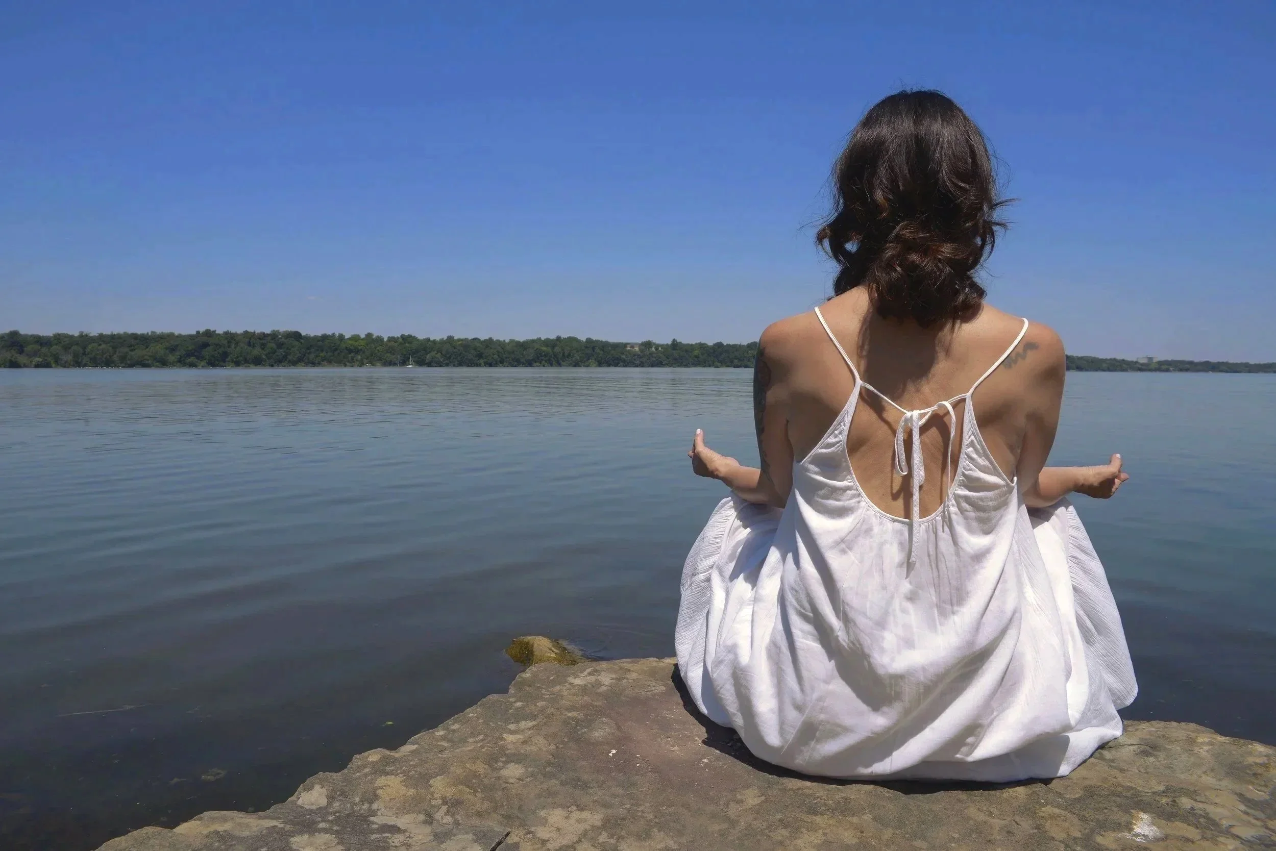 Woman in a white dress sitting on rocks by a calm lake during daytime, facing away from the camera.