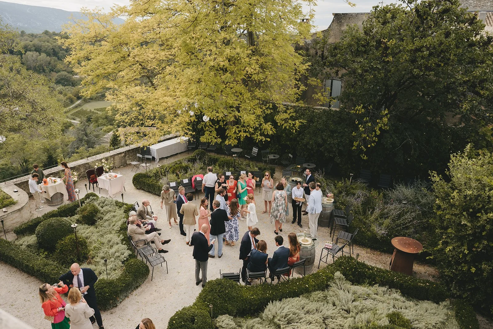 People attending an outdoor celebration or wedding reception on a terrace with lush trees and mountains in the background.
