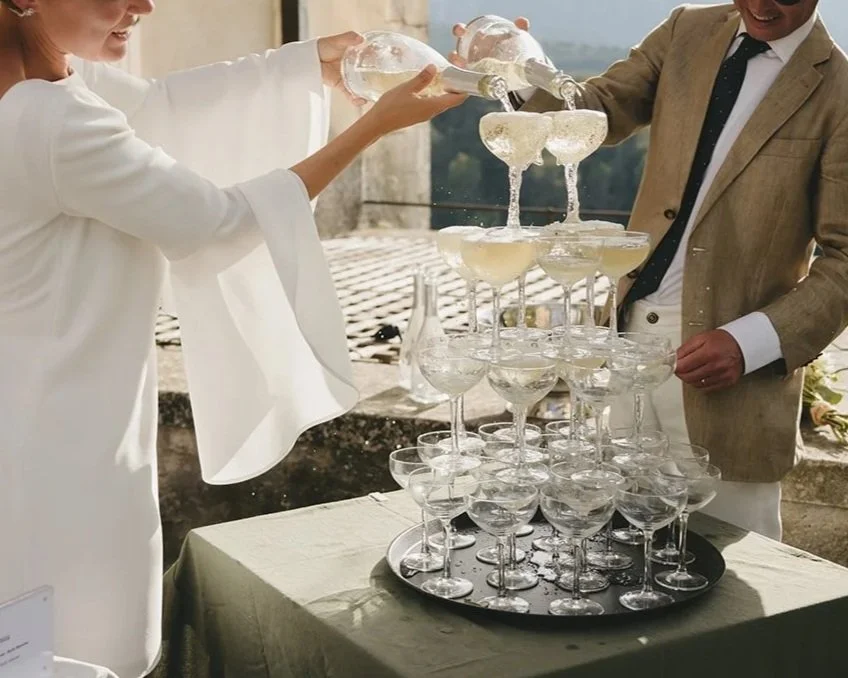 Two people are filling champagne glasses to create a layered champagne tower at an outdoor celebration event.