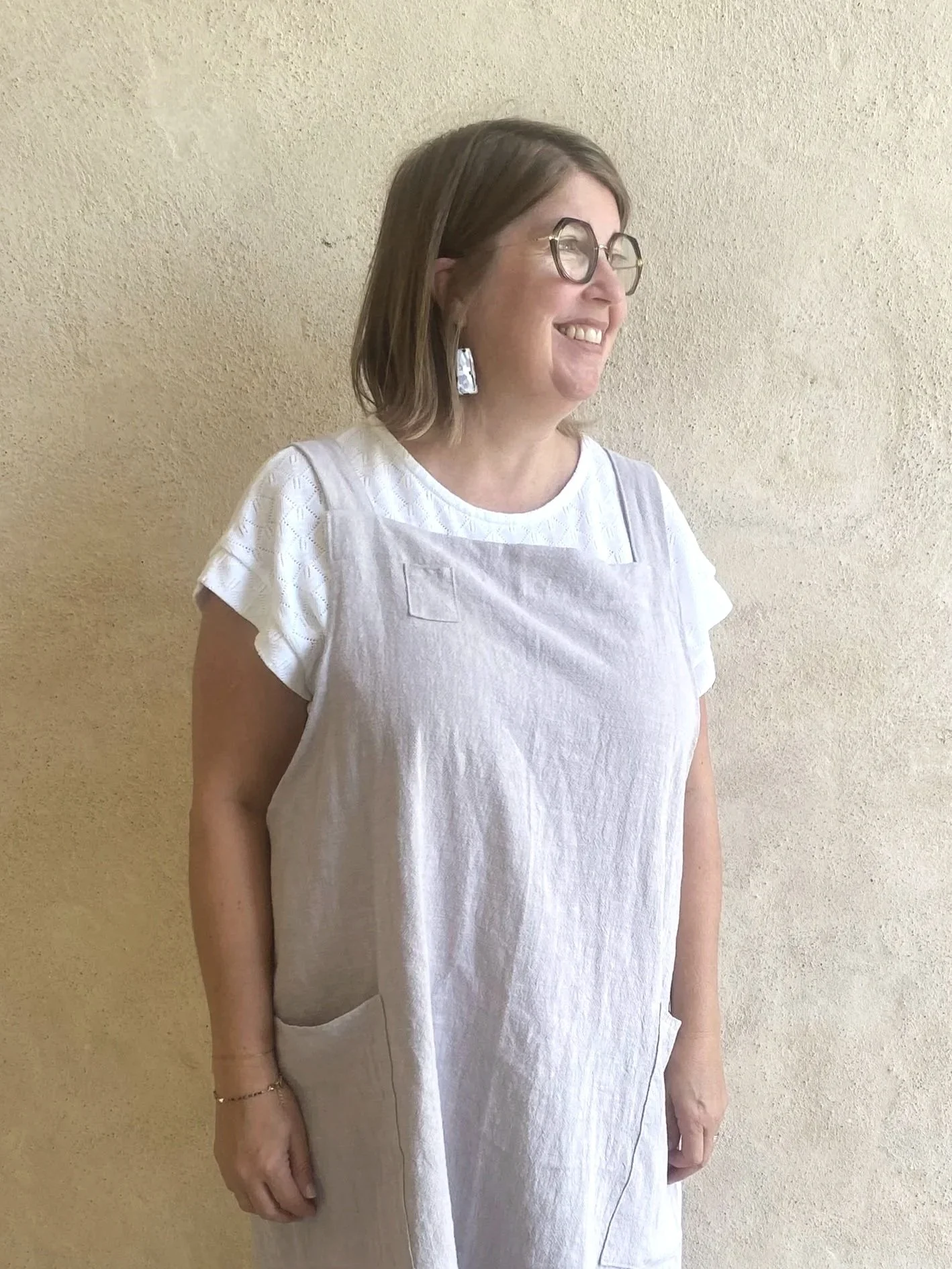 A woman with shoulder-length brown hair, wearing glasses, earrings, a white dress, and an apron, smiling and standing against a textured beige wall.