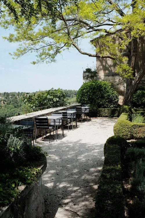 Outdoor seating area with black and white chairs and tables on a gravel path, surrounded by lush greenery and trees, overlooking a scenic view.
