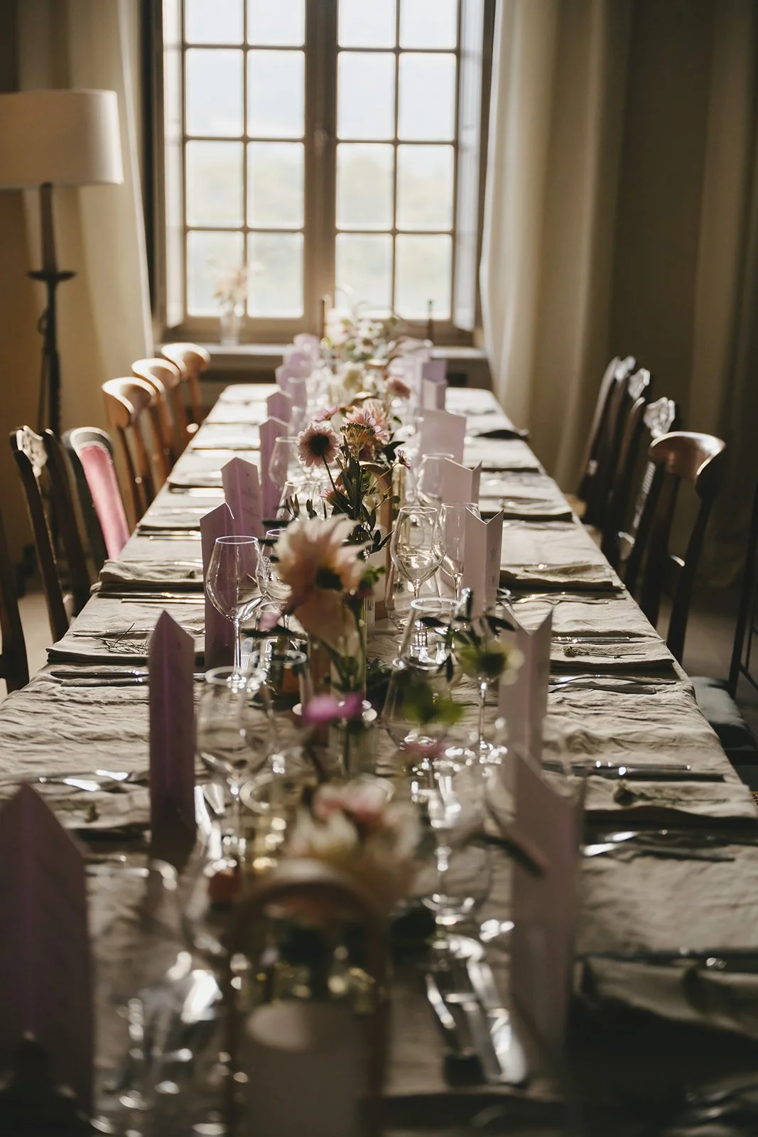 A long dining table set with flowers, glassware, and tableware in a room with large windows.