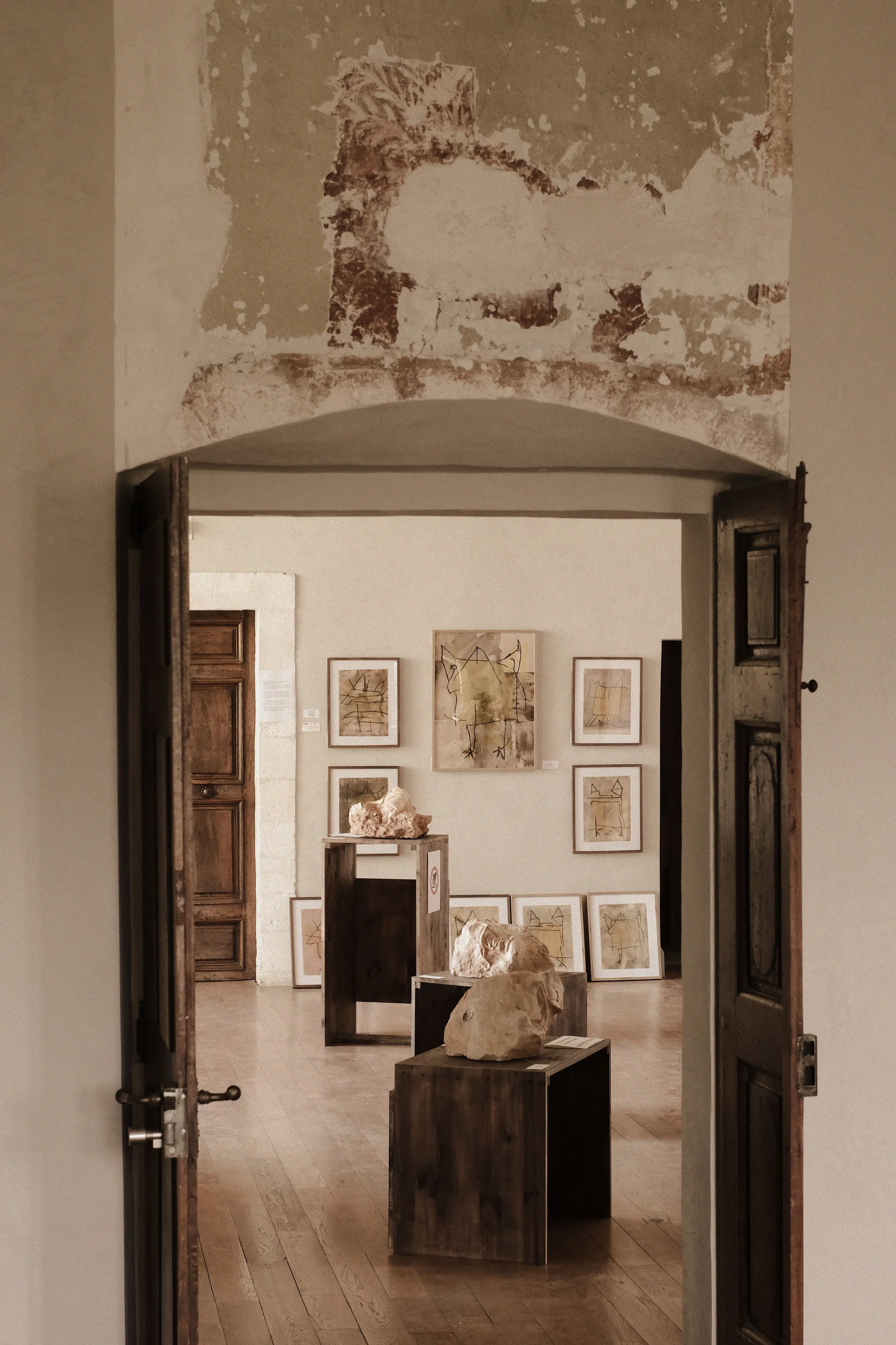 An art gallery interior seen through an open doorway, with framed abstract artwork on the wall and sculptures displayed on wooden pedestals in the center.