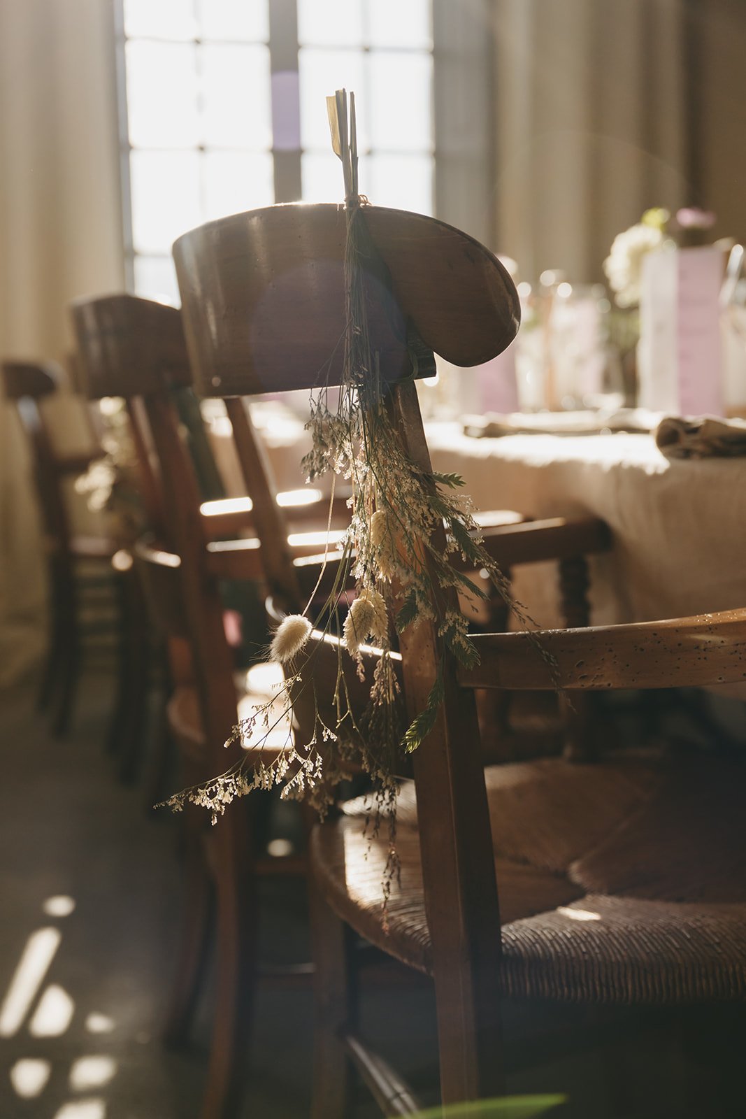 A wooden chair decorated with dried flowers hanging from the top, with sunlight streaming through a window in the background, suggesting a rustic or vintage setting.