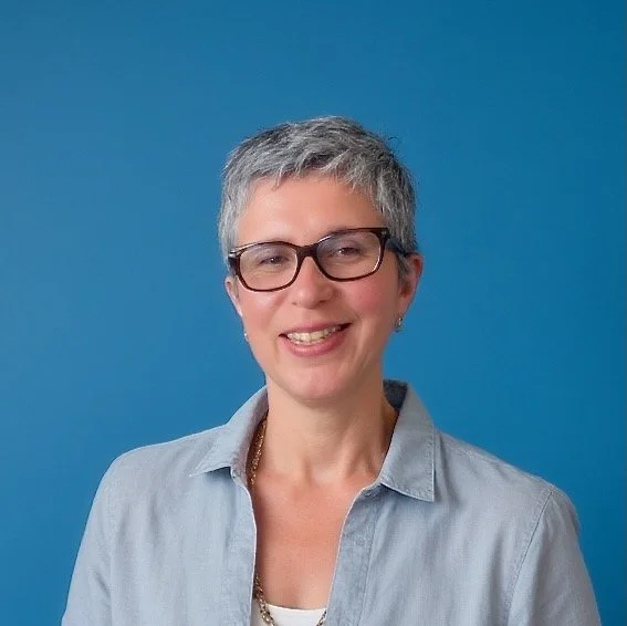 A smiling woman with short gray hair, wearing glasses and a light gray collared shirt, standing against a blue background.