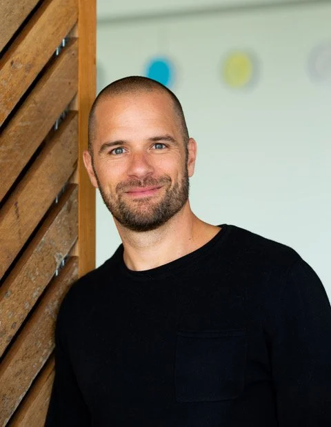 Man with a beard smiling, standing next to a wooden slat wall, in front of a white background with colorful circles.