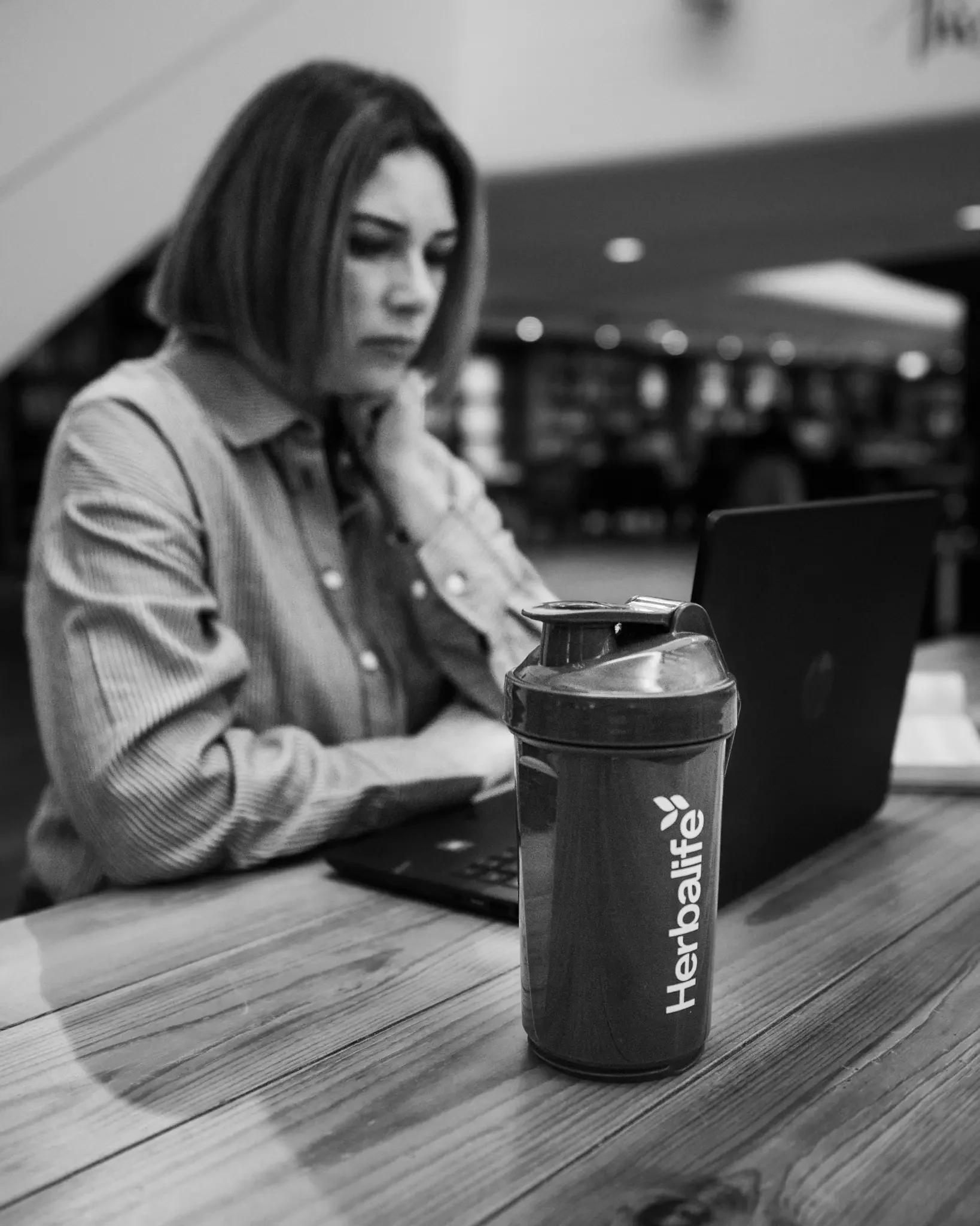 A woman sitting at a table in a cafe, working on a laptop, with a herbafit branded water bottle in the foreground.