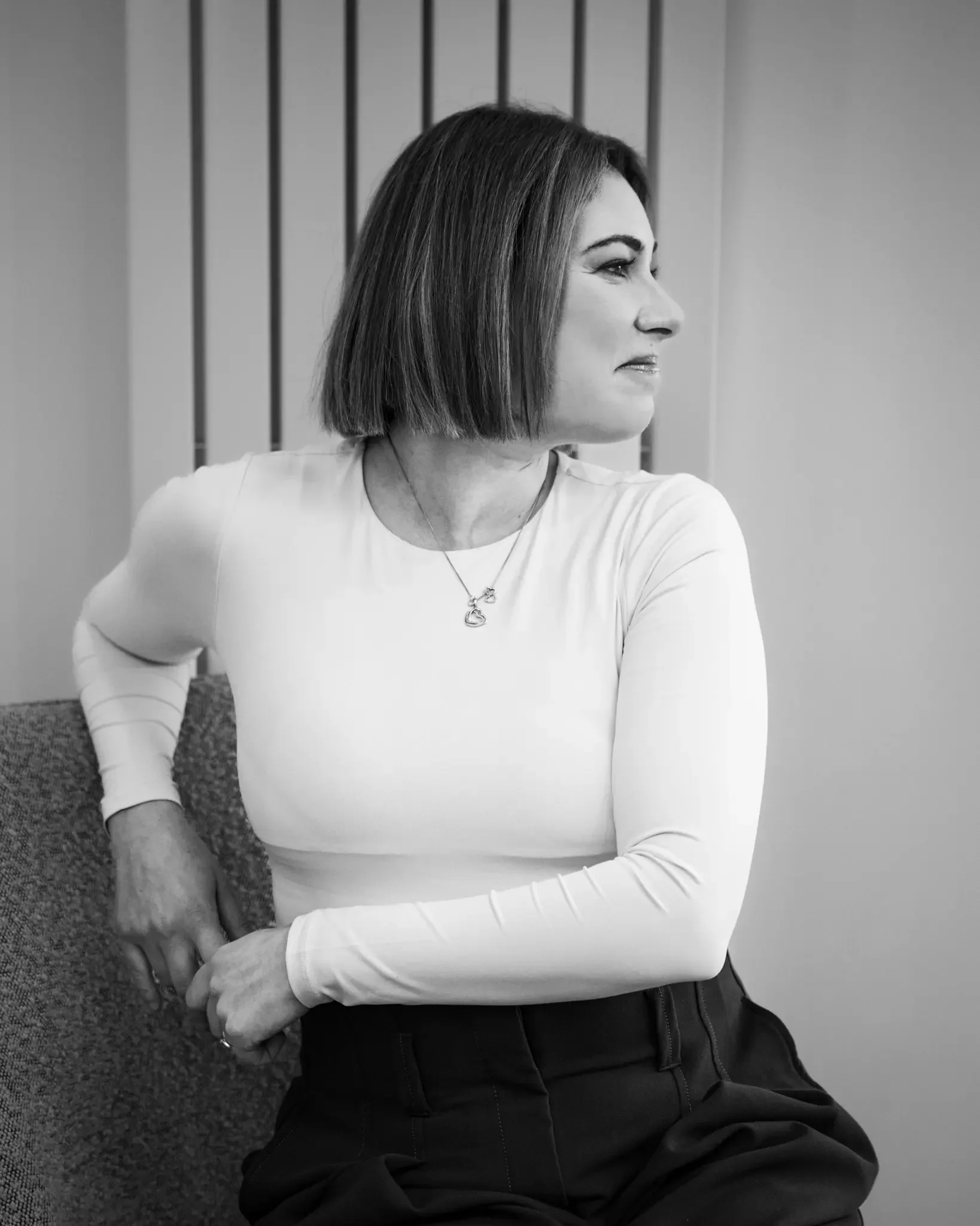A woman with shoulder-length hair sits on a textured chair, wearing a white long-sleeve top and dark pants, looking to her right.