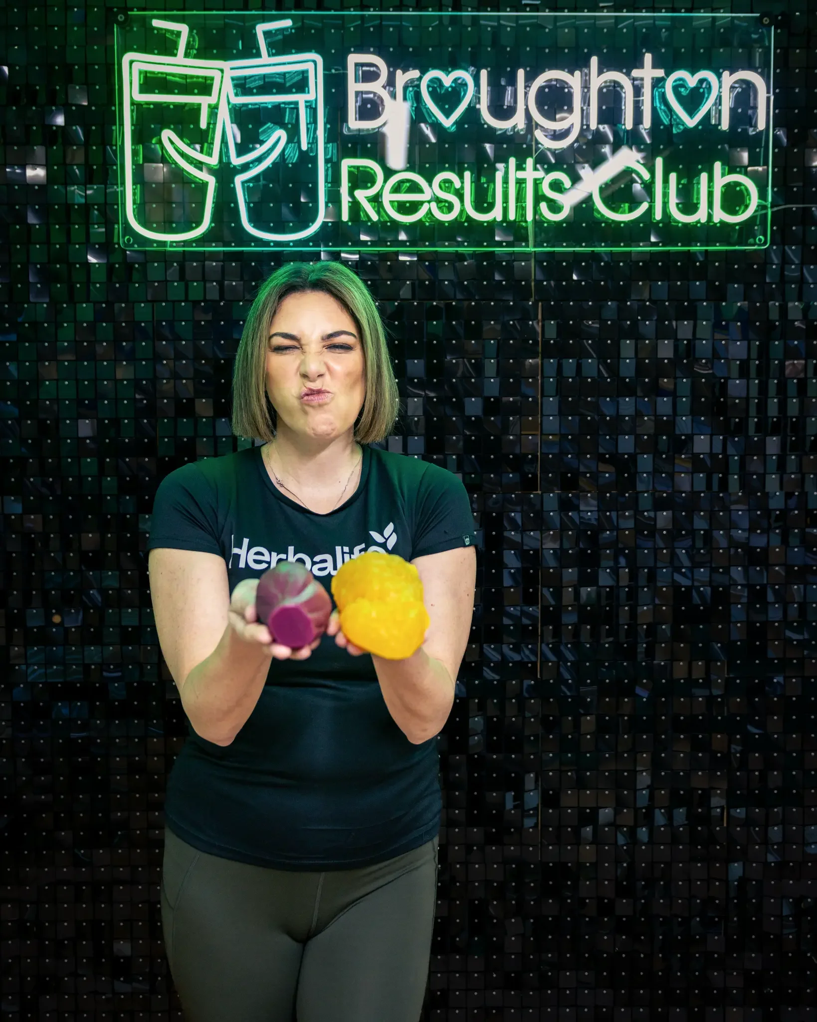 A woman wearing a Herbalife shirt holding a dragon fruit and a yellow passion fruit in front of a black textured wall with a neon sign that reads 'Brughton Results Club' and includes two drink glasses with straws.