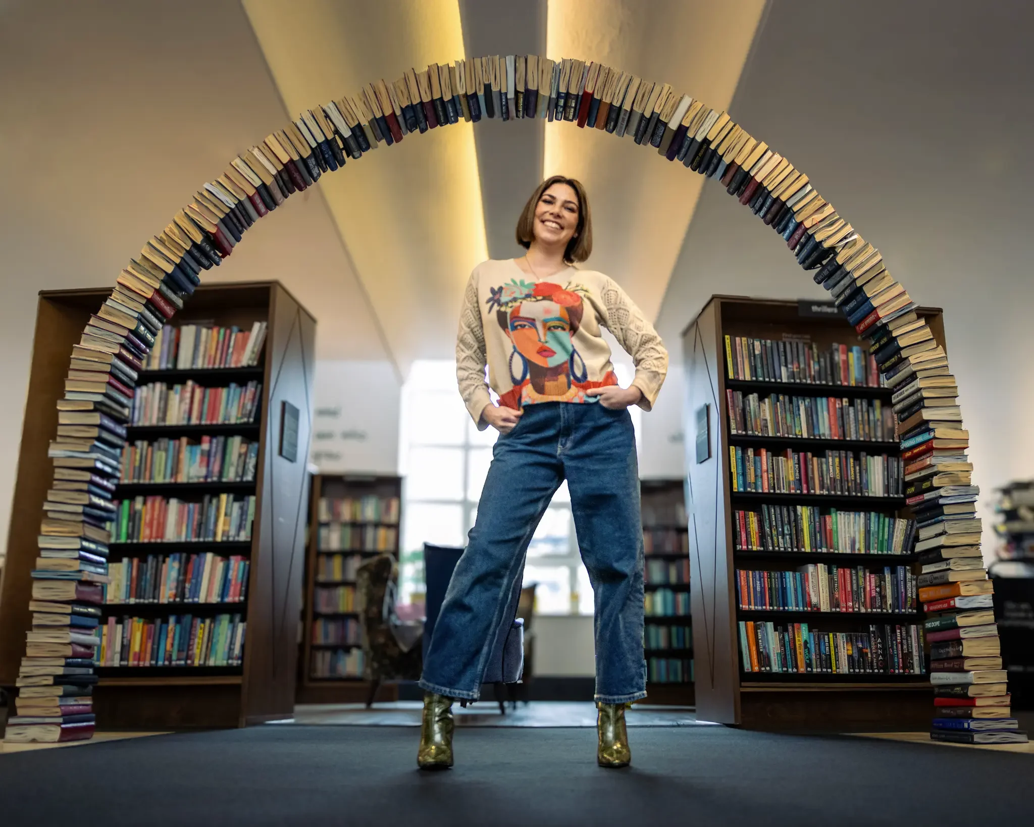A woman standing in a library under a large arch made of books, wearing a colorful sweater, jeans, and metallic boots, smiling.