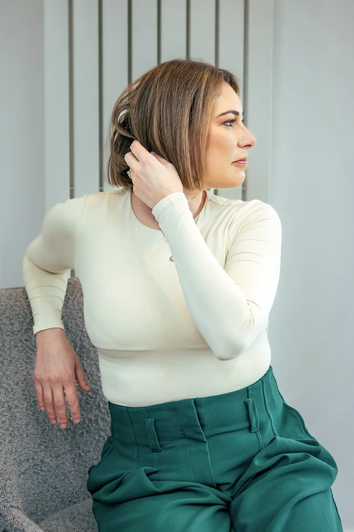 A woman with shoulder-length brown hair sitting on a gray chair, wearing a cream-colored long sleeve shirt and green pants, with her left hand resting on the chair and her right hand touching her hair, looking to her right.