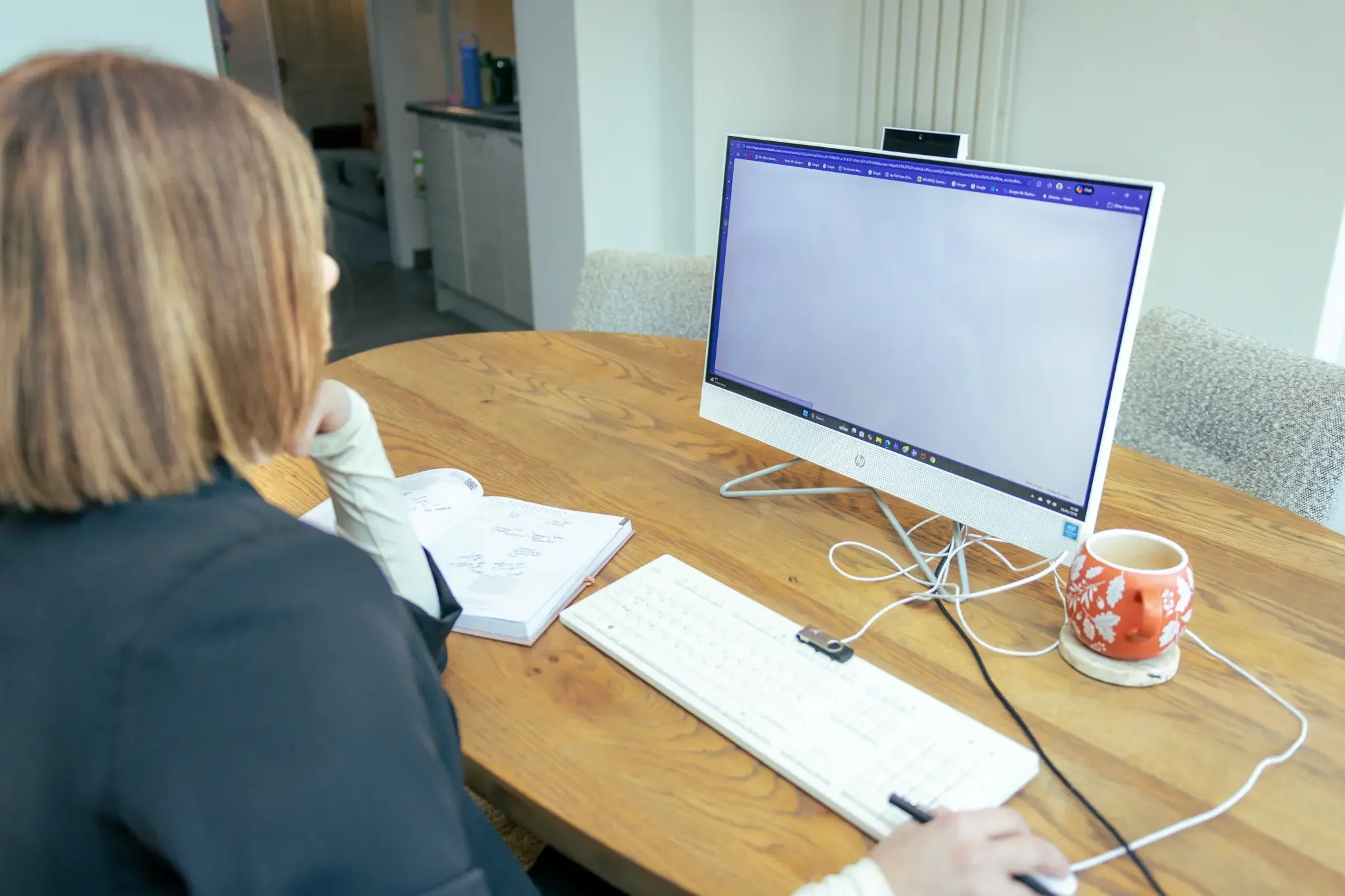 A woman with shoulder-length hair sitting at a wooden table, working on a computer with a large monitor, open notebooks, and a cup of coffee.