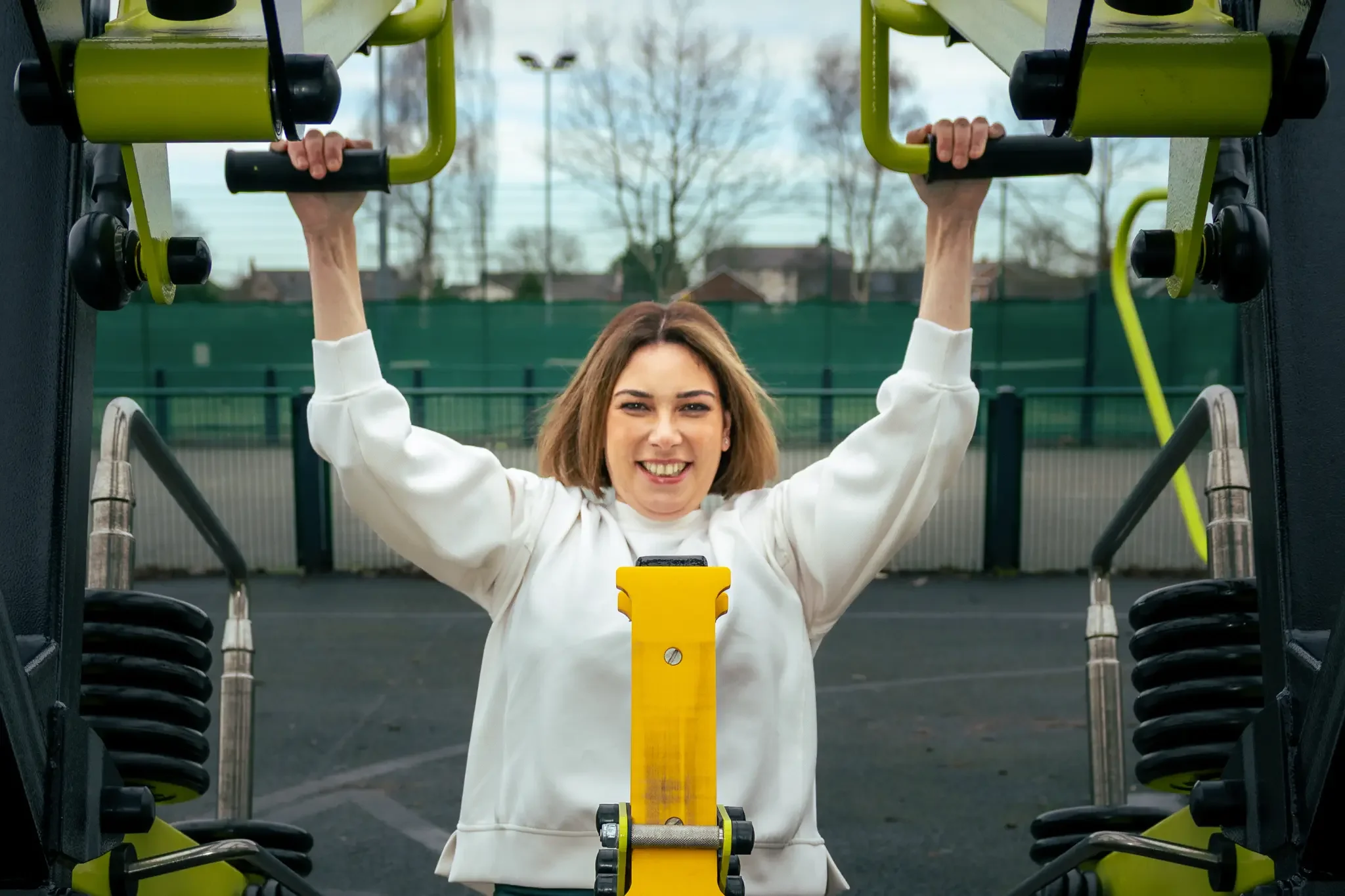 A young woman in a white hoodie smiling and hanging from outdoor gym equipment with green and black structures and a sports field in the background.