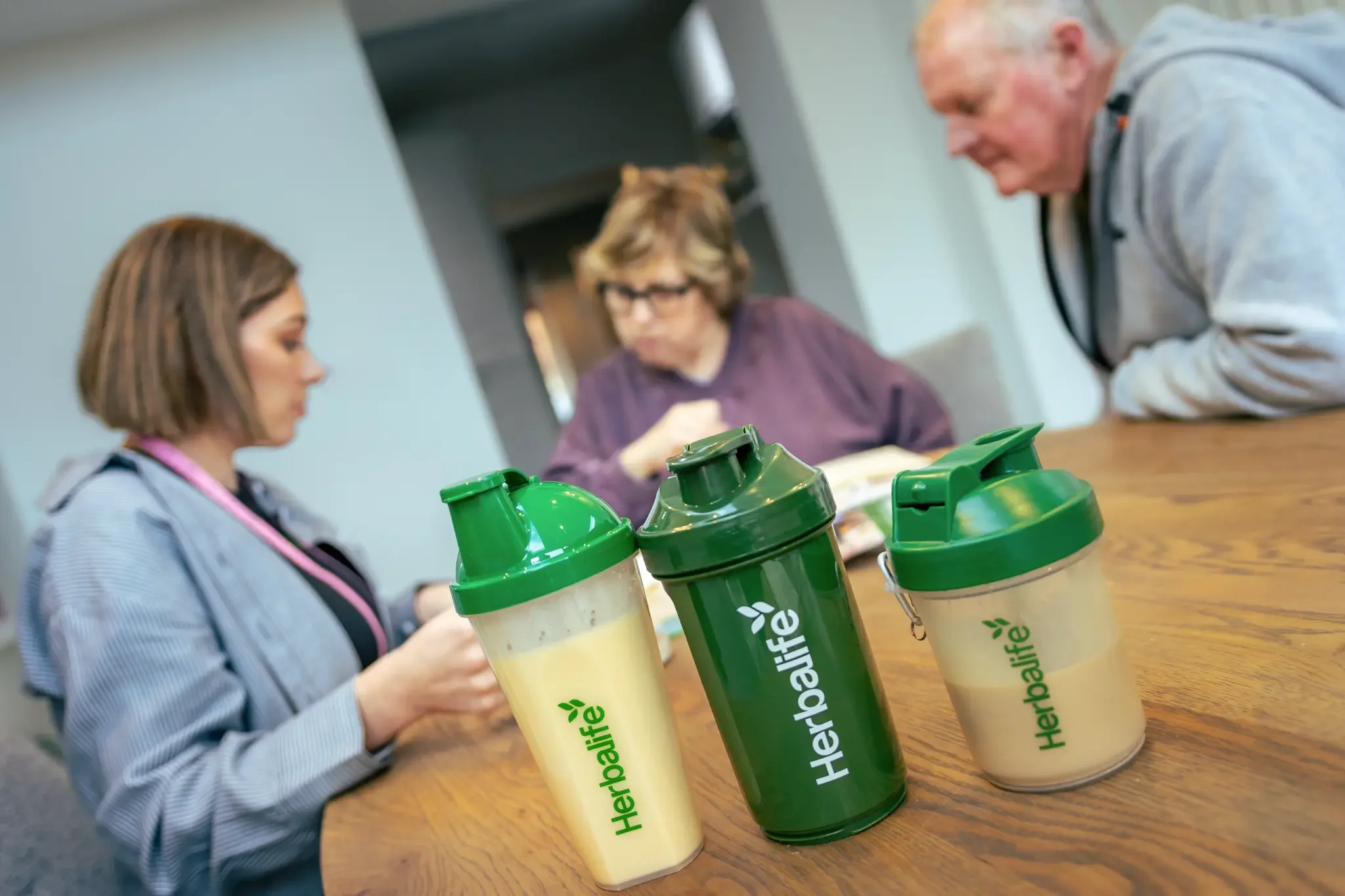 Three Herbalife protein shake containers on a wooden table with three people in the background, one woman and two men, looking at their phones and talking.
