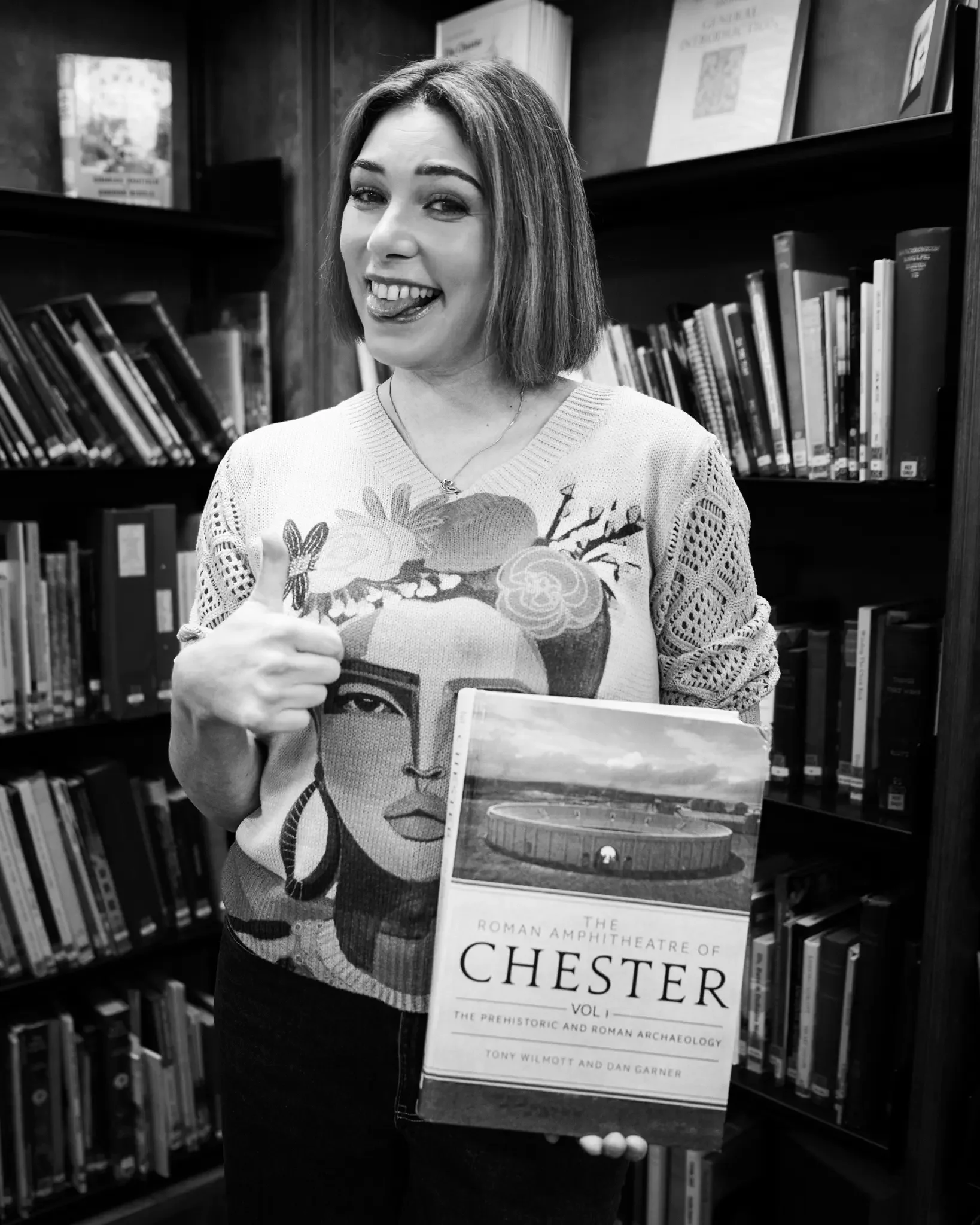 A woman holding a book titled 'The Roman Amphitheatre of Chester' in a library, with bookshelves in the background, giving a thumbs-up and sticking her tongue out.