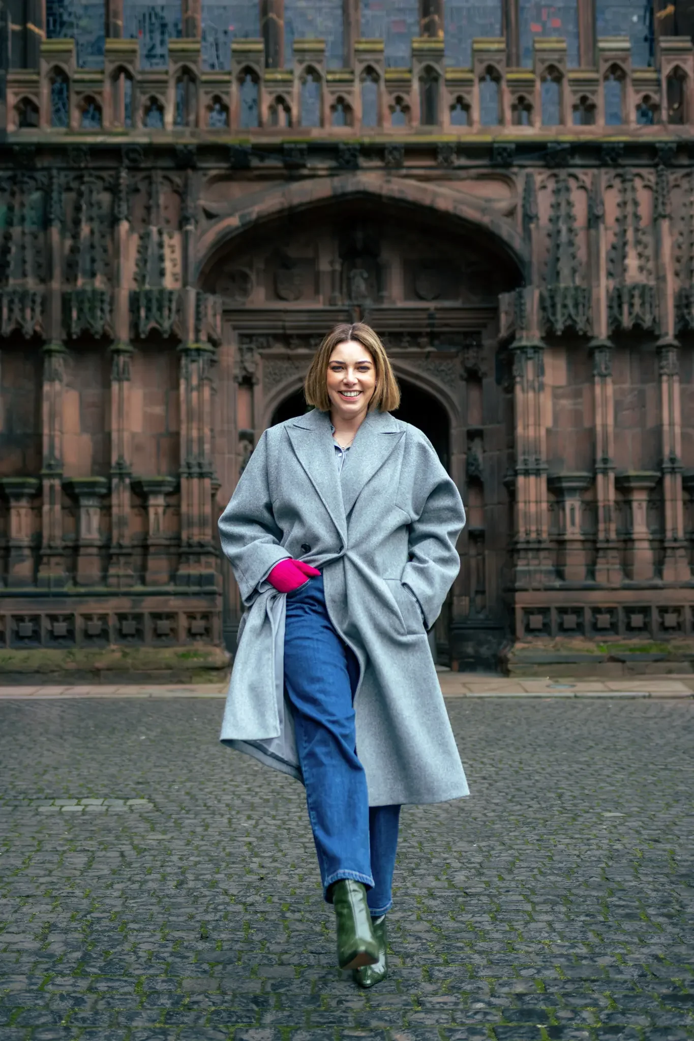 A woman with short brown hair smiling while walking outdoors on a cobblestone street in front of a historic building with elaborate gothic architecture, wearing a long gray coat, blue jeans, green boots, and pink gloves.