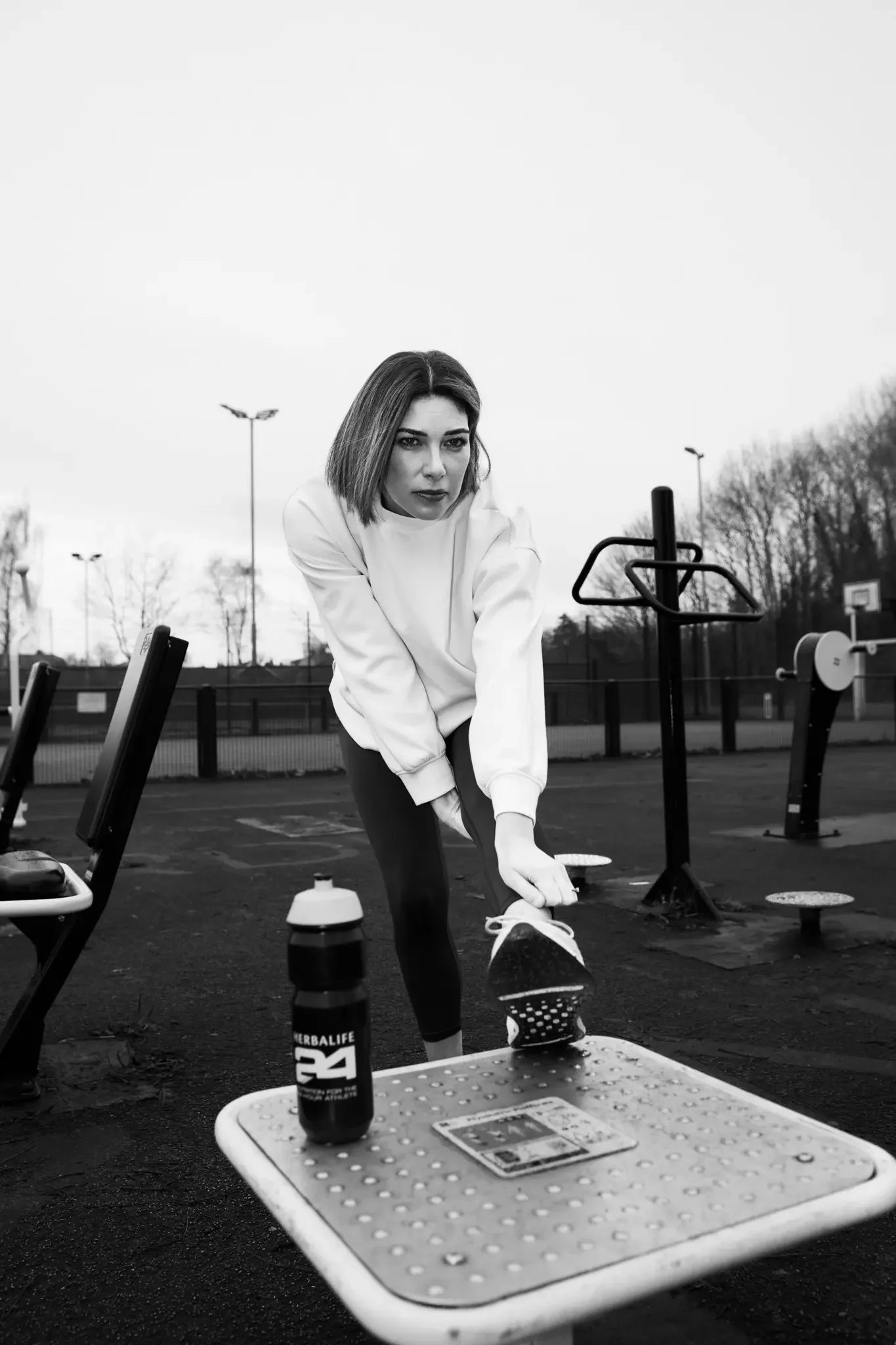 A woman in athletic clothing stretching outdoors at a park or playground, with exercise equipment in the background and a water bottle on a nearby table.