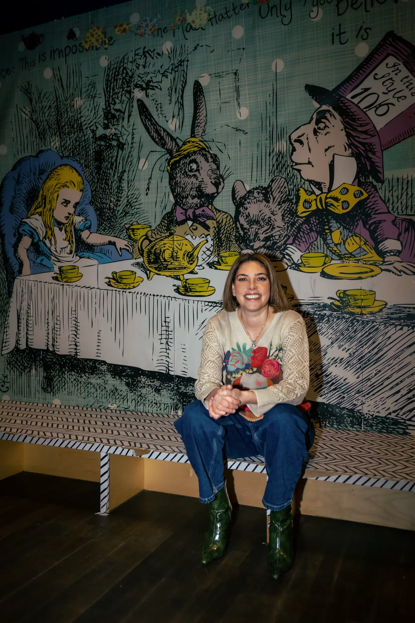 A woman sitting in front of a large Alice in Wonderland-themed mural depicting the Mad Hatter's tea party with a woman, rabbit, mouse, and a cat, all enjoying tea.