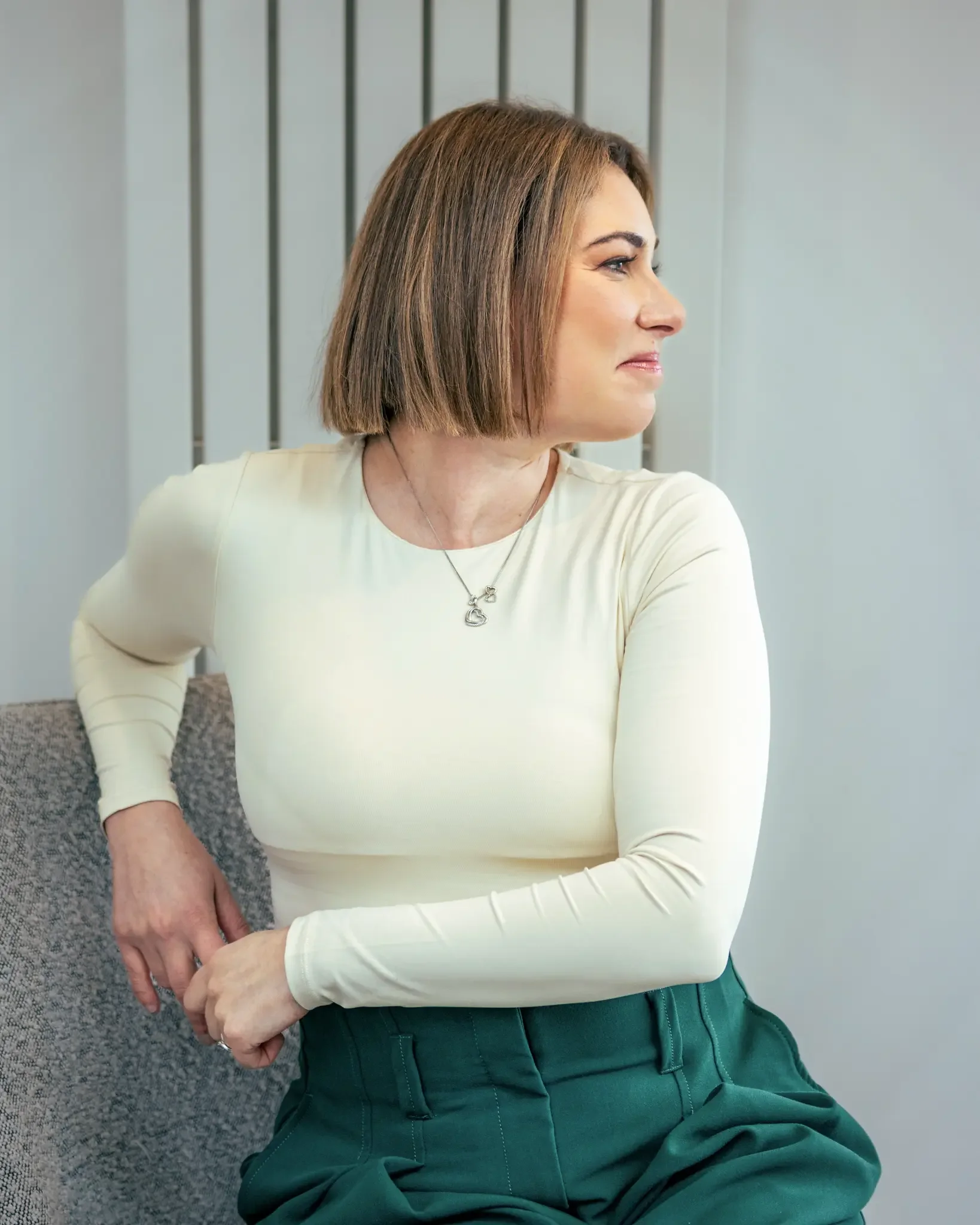 Woman sitting against a gray background, wearing a cream long-sleeve top and green pants, looking to her right.