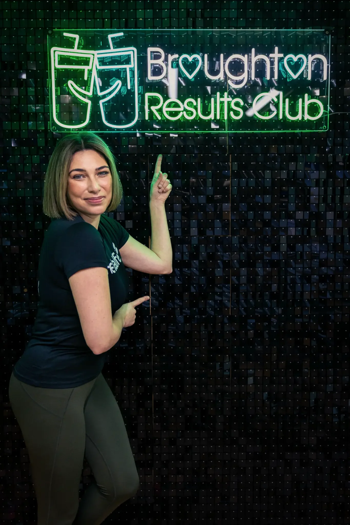 Woman pointing at a neon sign that reads 'Broughton Results Club' with an icon of two drinks, against a black tiled background.
